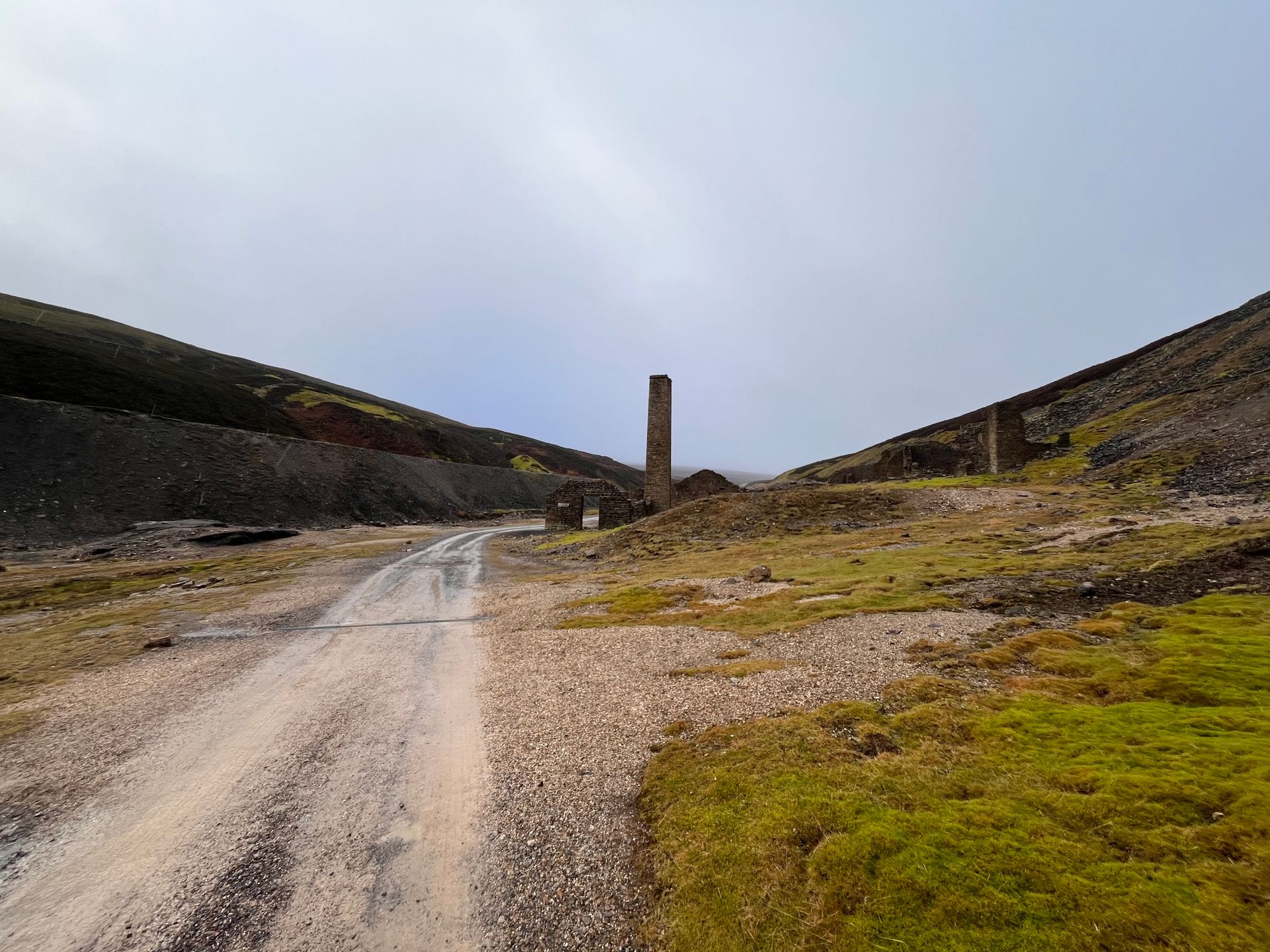 A gravel path leads toward the tall, stone chimney stack of an old mining ruin in a barren, hilly landscape.