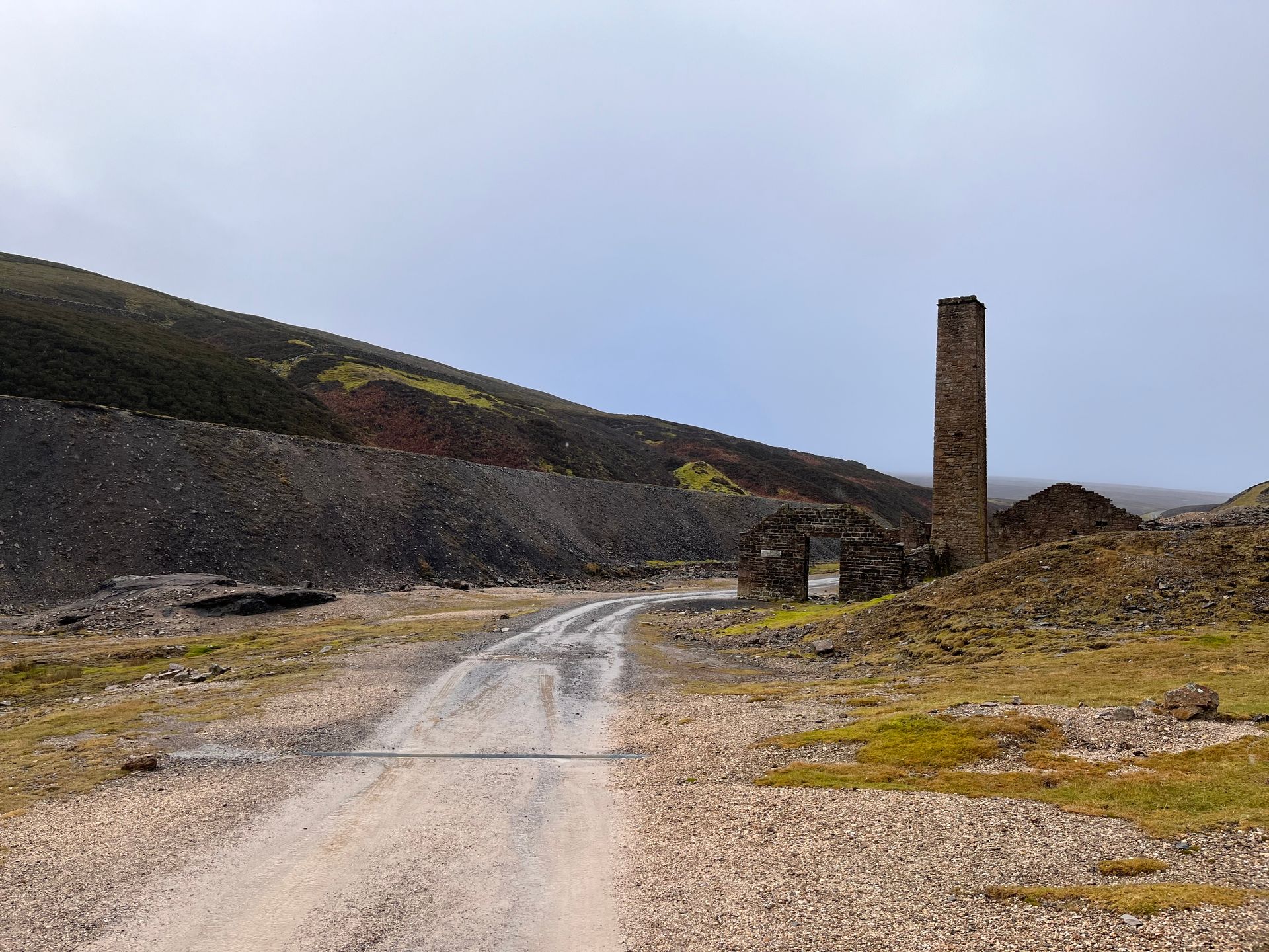 A gravel track leads toward a weathered stone chimney and mine ruins nestled against a dark, rocky hillside.