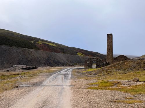 A gravel road leads toward a stone chimney stack and ruins set in a stark, hilly landscape under a grey sky.