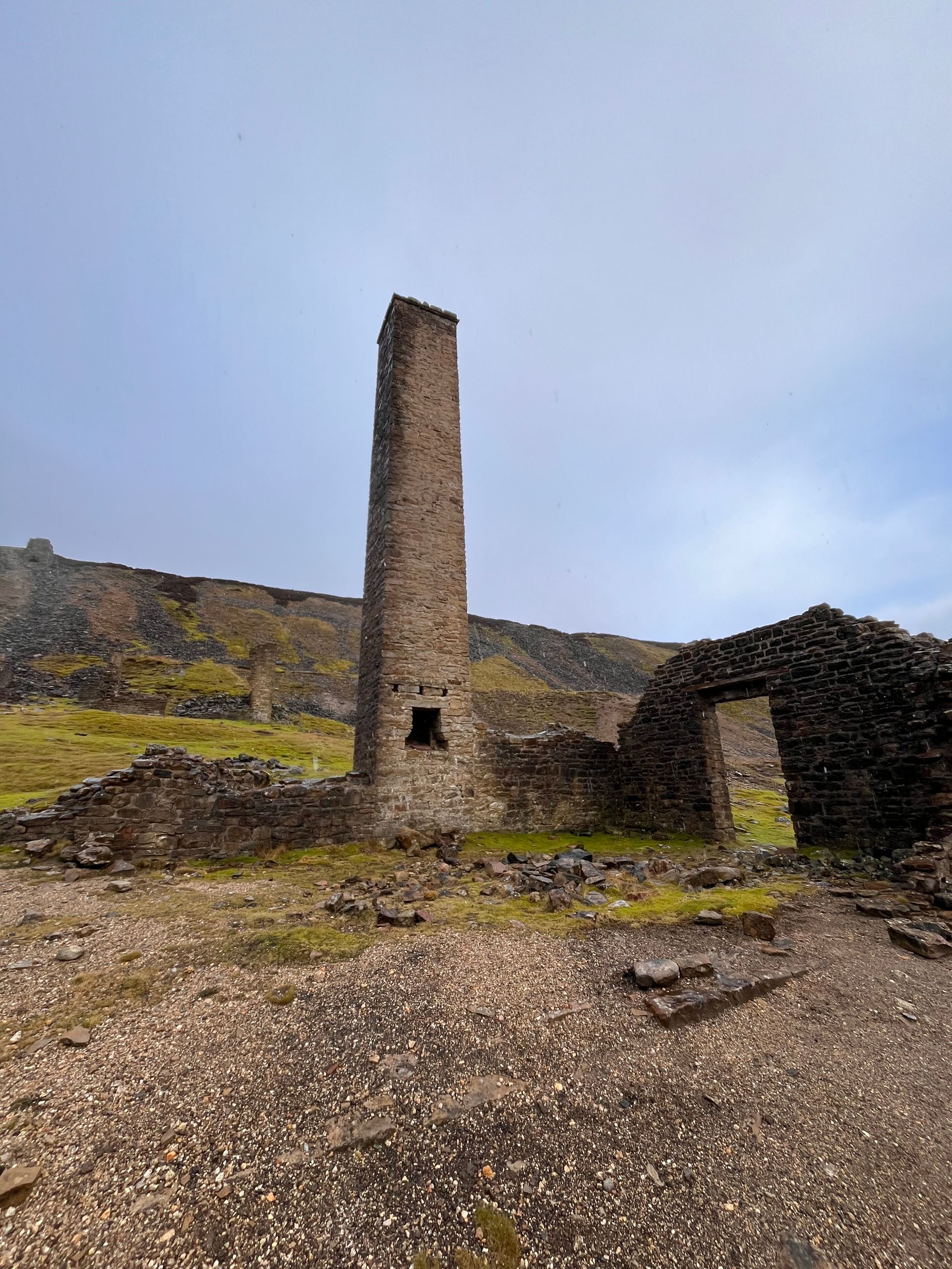 Tall, stone chimney remains standing among ruined walls in a rocky, grassy landscape under a cloudy sky.