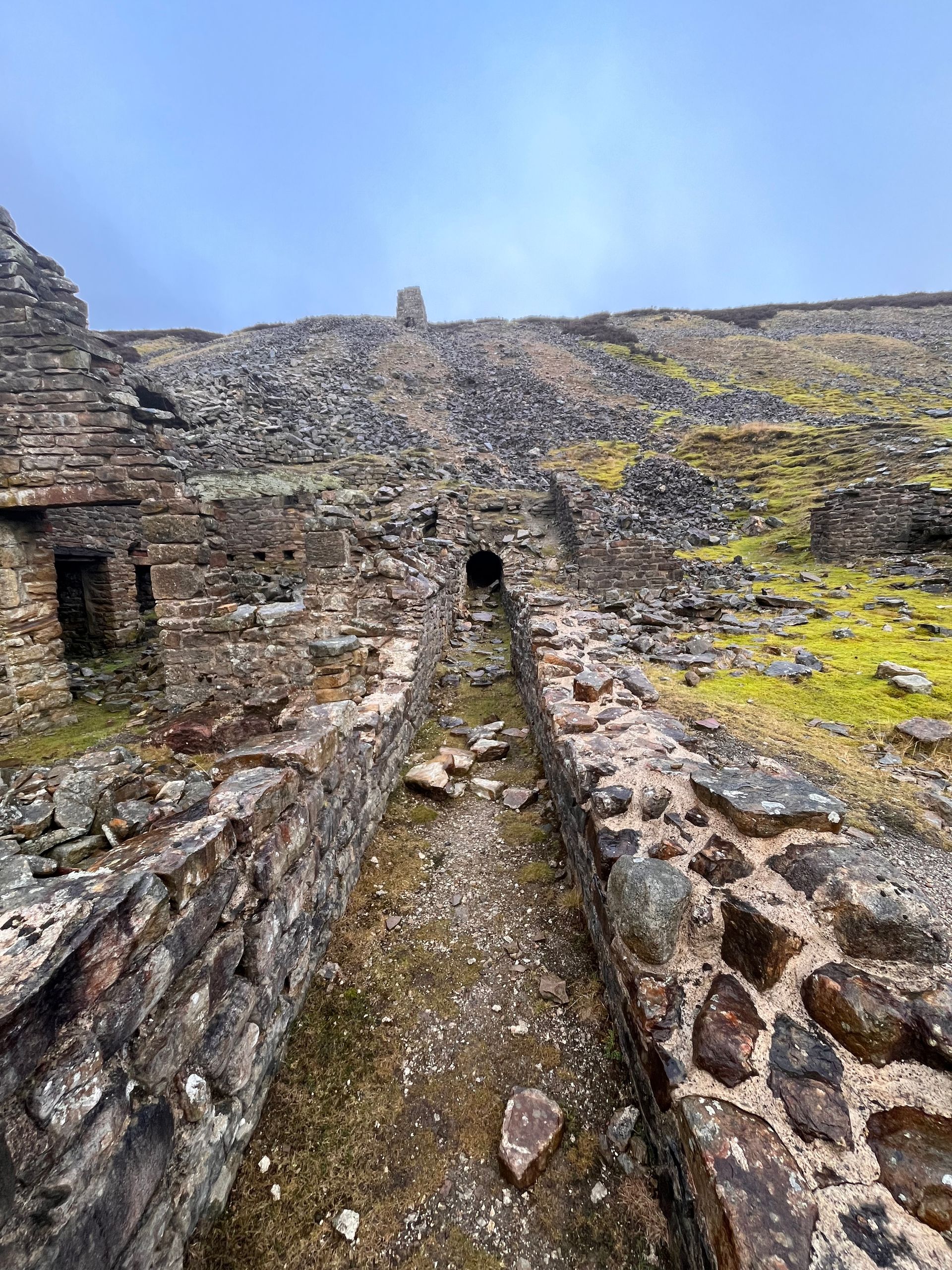 A narrow stone-walled channel leads toward ruins under a cloudy sky in a rocky, mossy landscape.