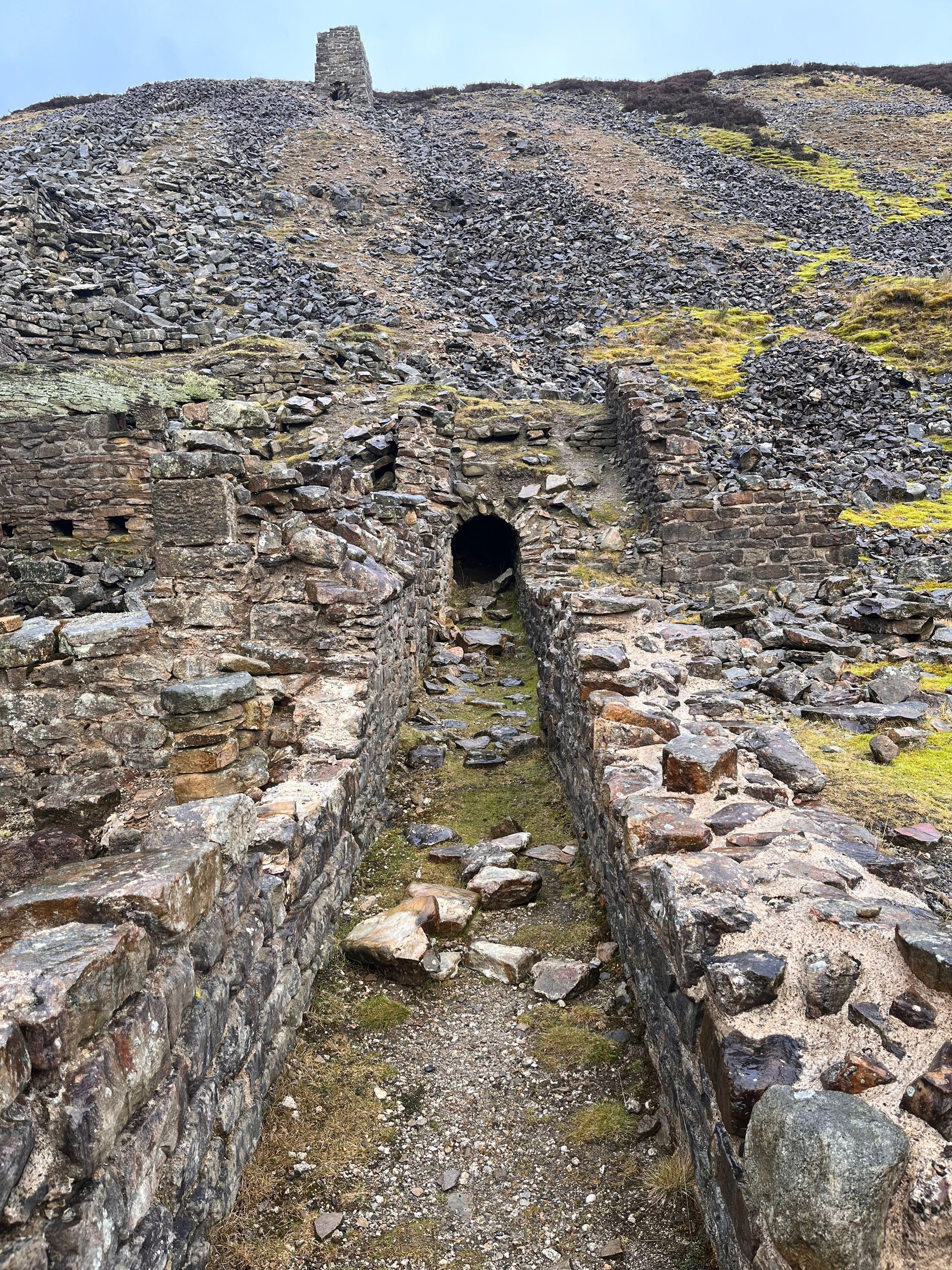 Stone ruins of an old mine entrance set against a steep, rocky hillside covered in grey rubble and patches of yellow moss.