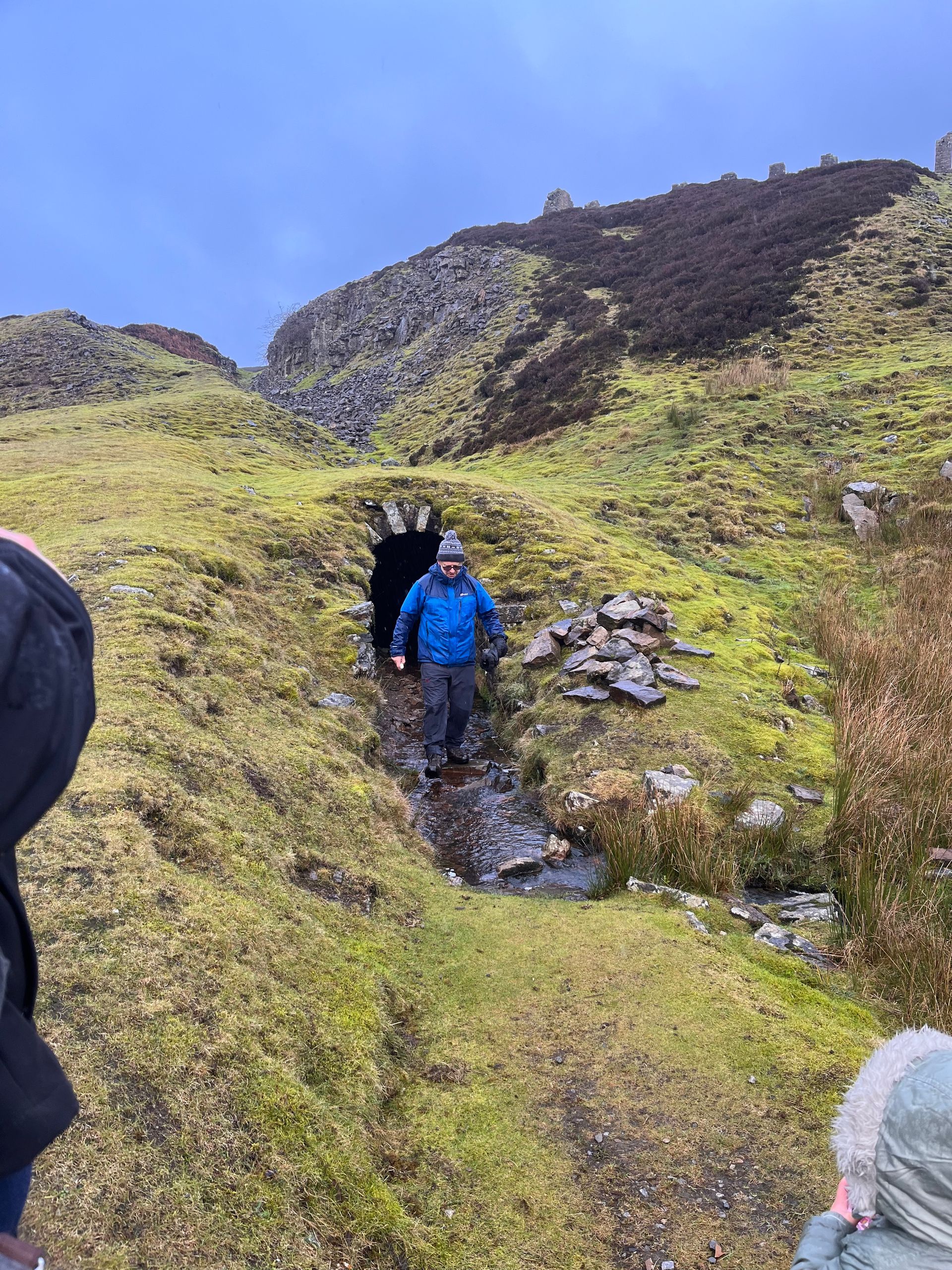 A person in a blue jacket walks out of a small, arched stone tunnel into a grassy, hilly landscape.