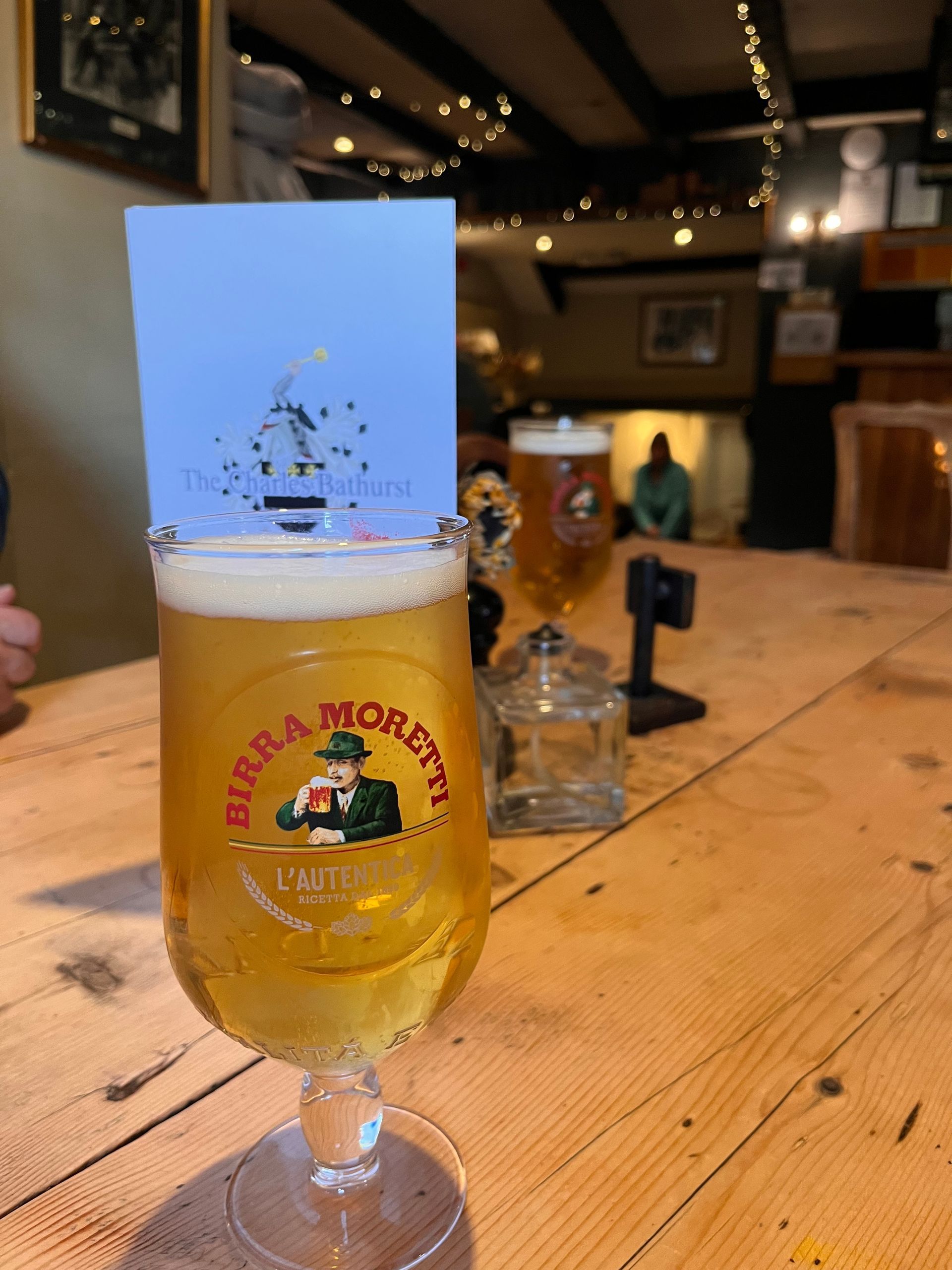 A glass of Birra Moretti beer sits on a rustic wooden table in a pub, with a small promotional card tucked behind it.