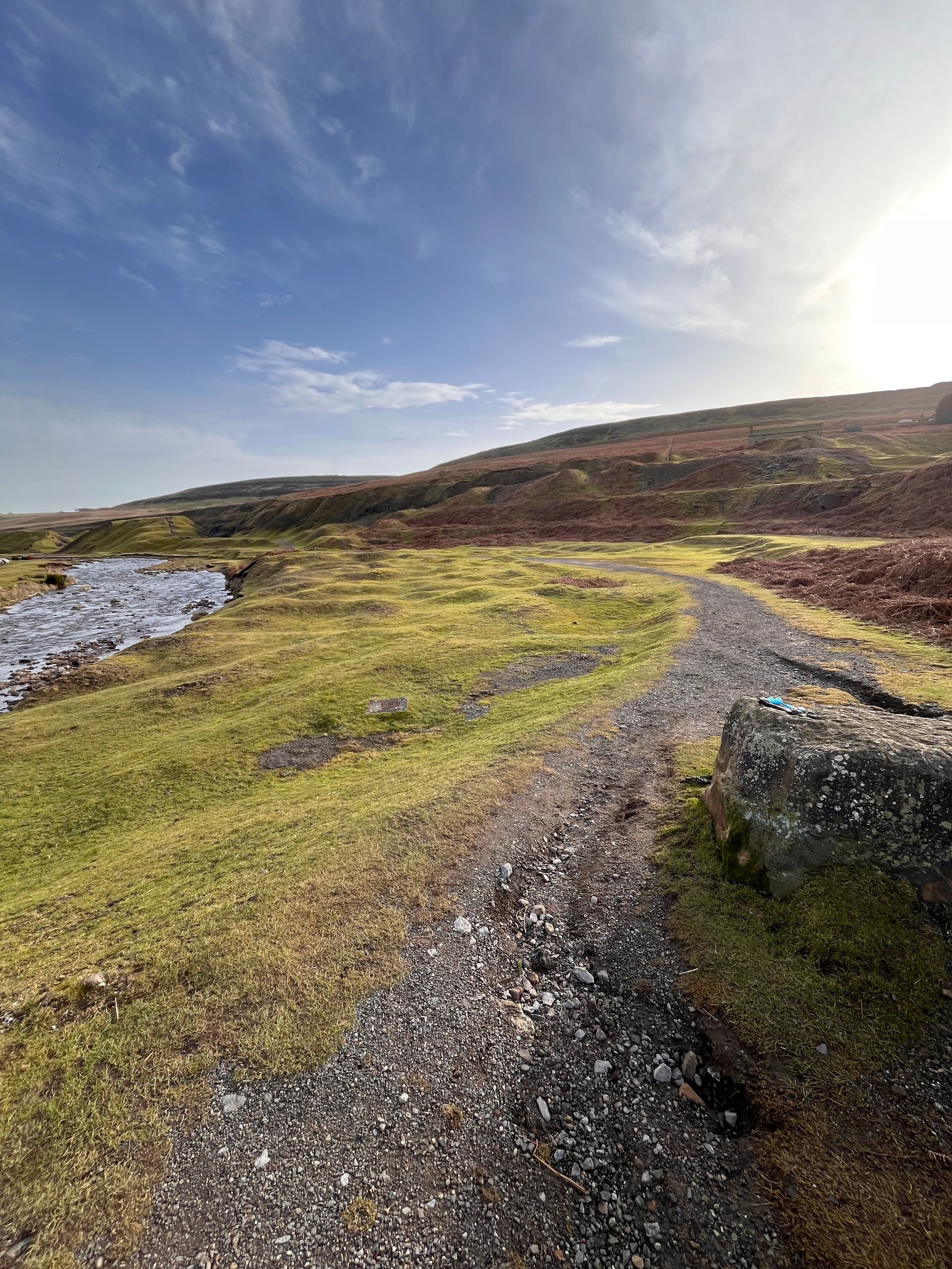 A dirt path winds through a grassy moorland landscape under a bright, sunny sky with low hills in the distance.