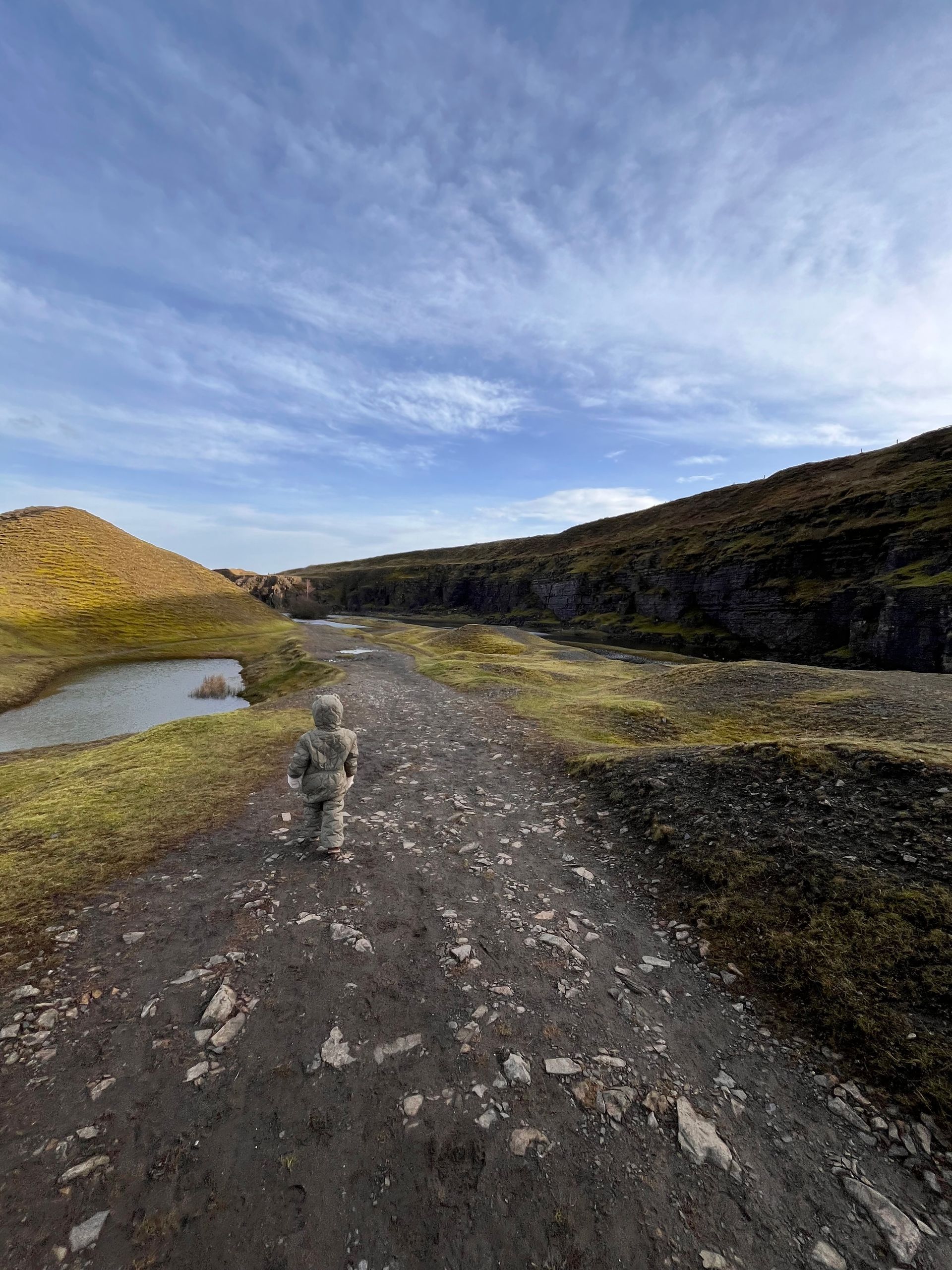 A person in a neutral-colored hooded jacket walks down a rocky, gravel path between grassy hills under a blue sky.