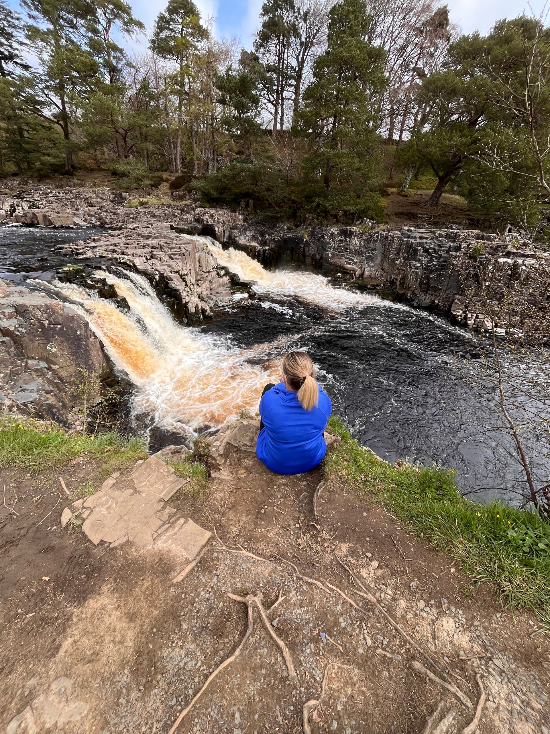 A person in a blue shirt sits on a grassy ledge overlooking a small, flowing river waterfall surrounded by trees.