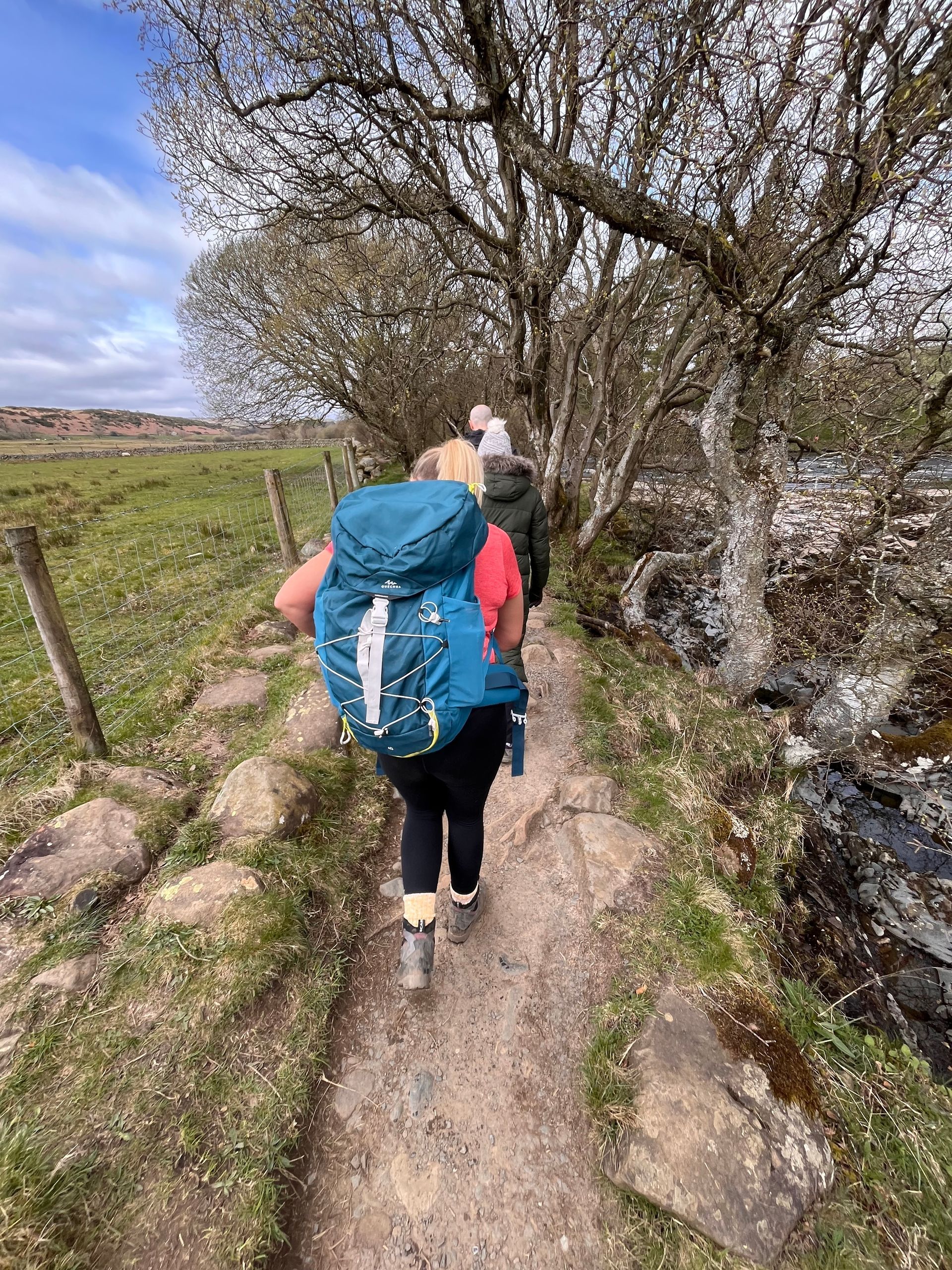 A person with a large blue hiking backpack walks along a dirt trail flanked by trees and a stone wall on a sunny day.