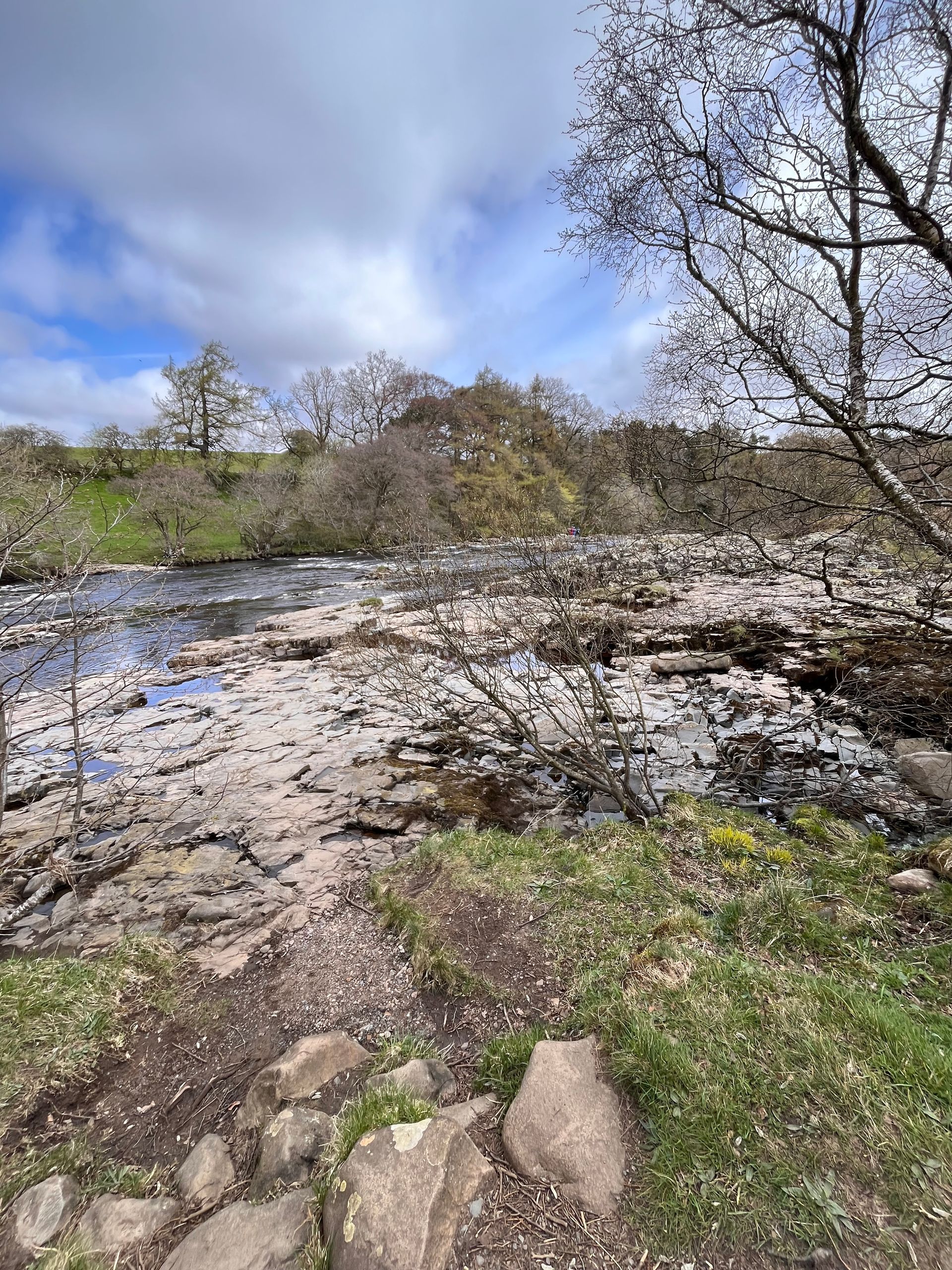 A rocky riverbank leading into a shallow stream with leafless trees under a cloudy blue sky.