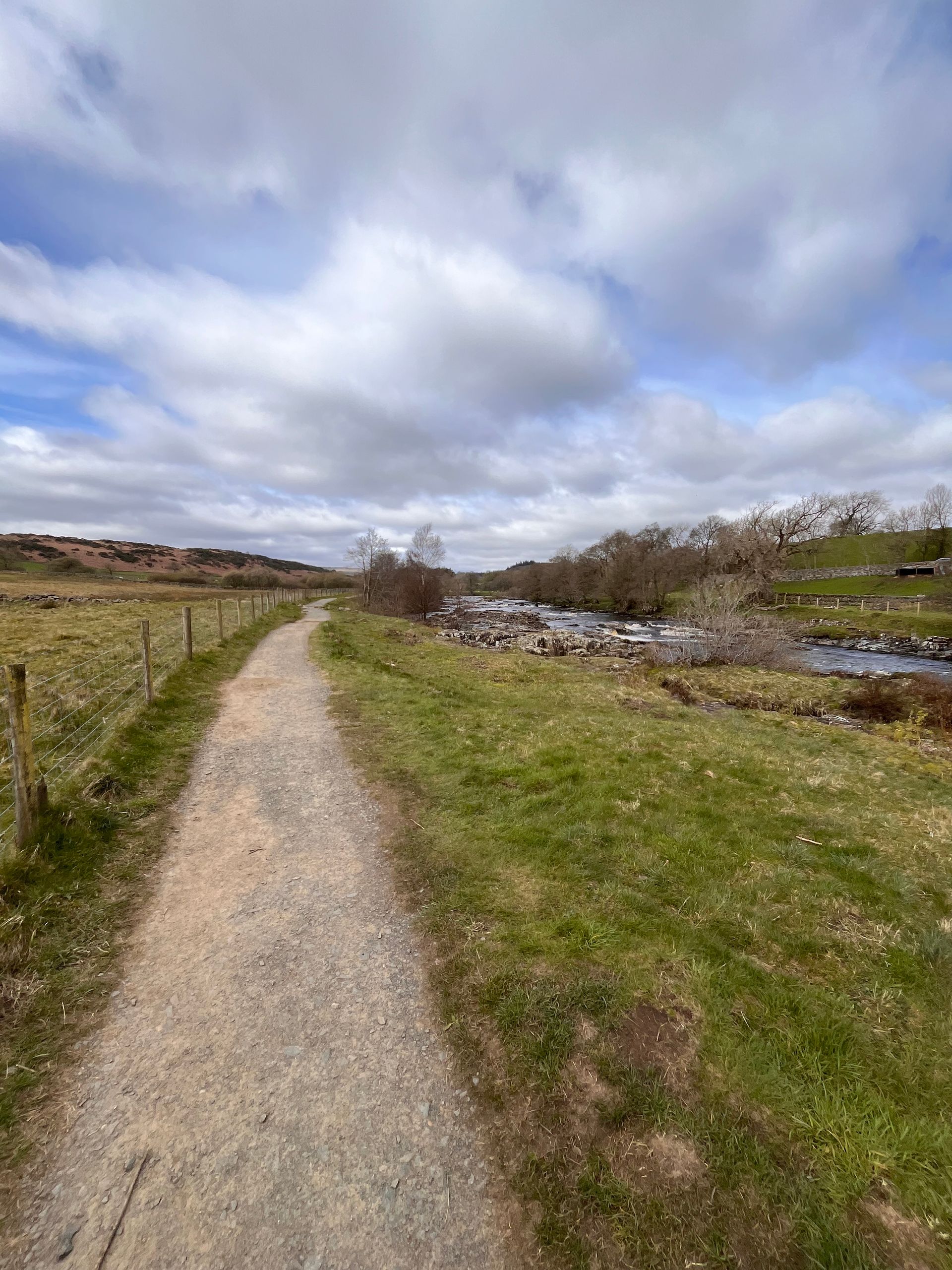 A dirt path winds through a grassy field toward a small stream under a cloudy, blue sky.