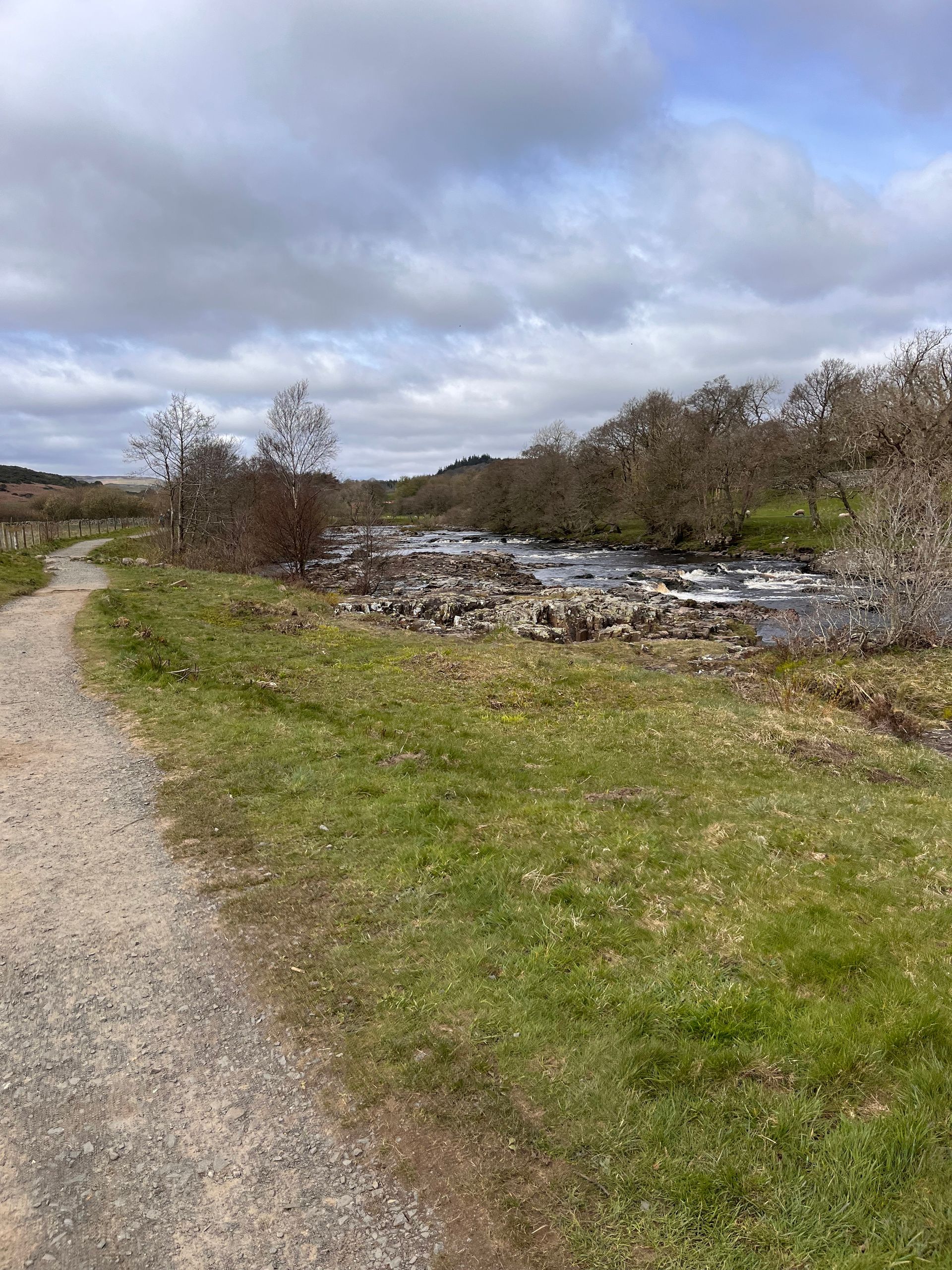 A gravel path winds along a rocky stream bordered by grass and bare trees under a cloudy sky.