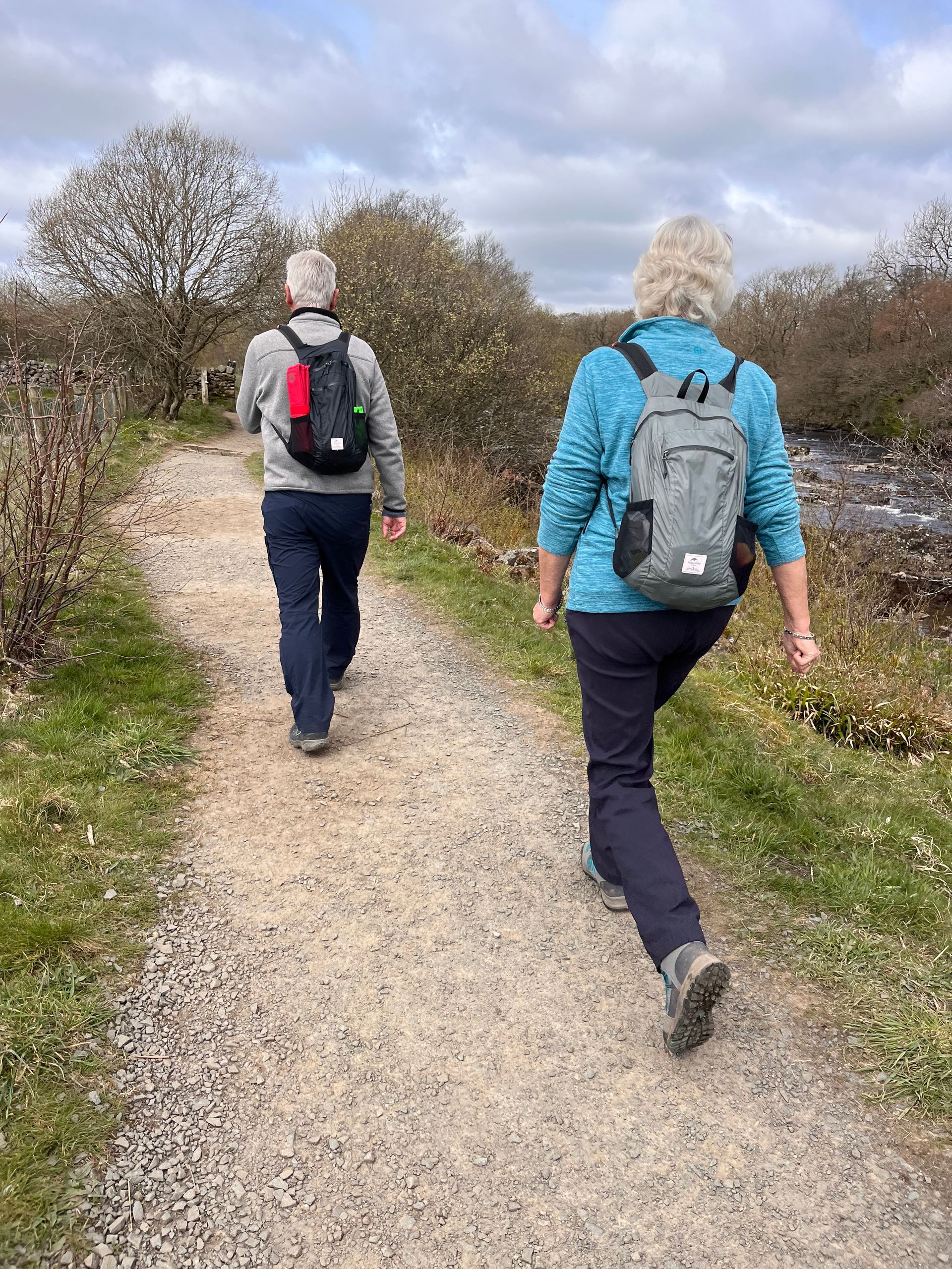 Two people with backpacks walking along a dirt path through a field with scattered trees under a cloudy sky.