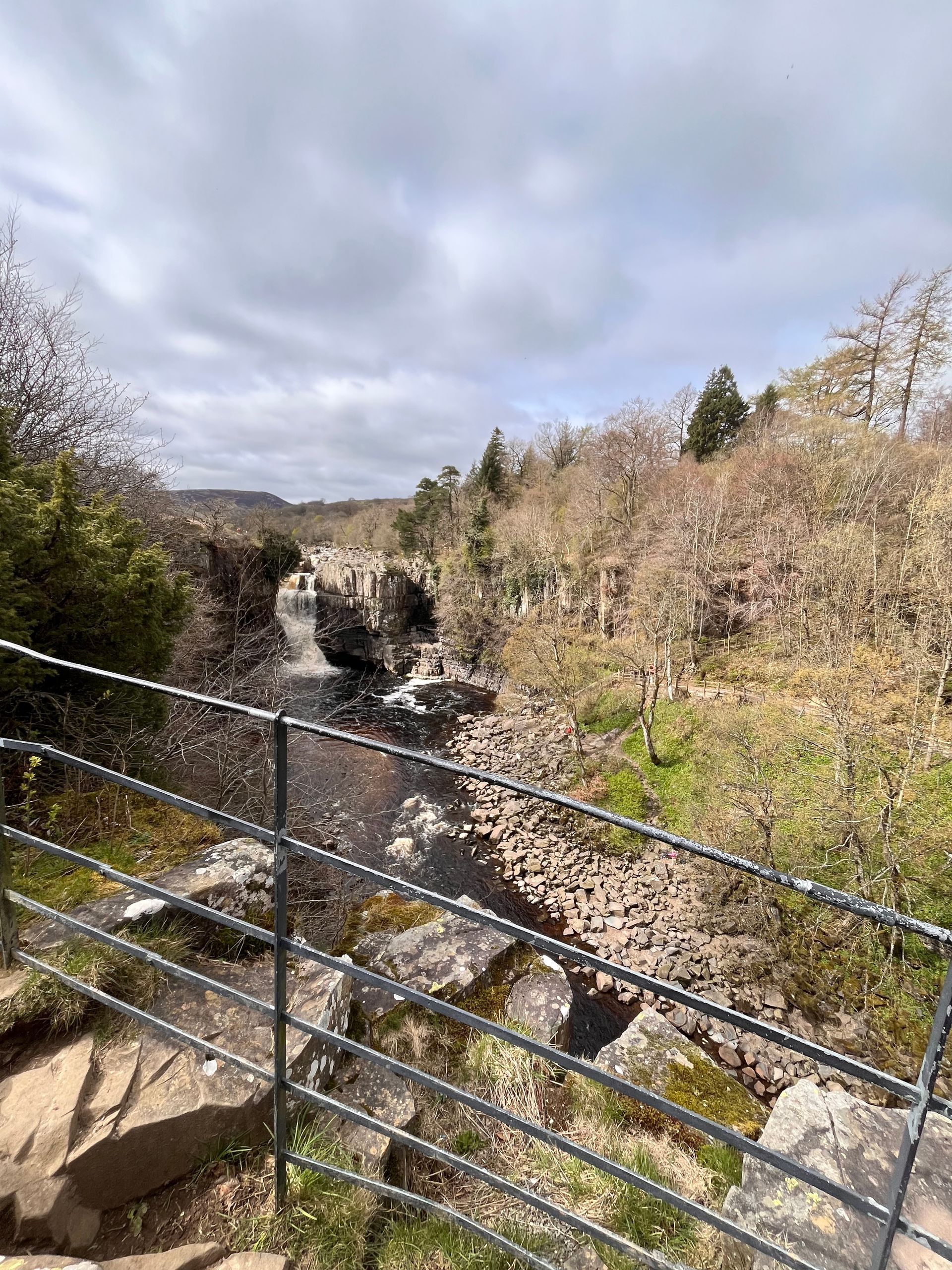 A small waterfall cascades into a rocky river bed surrounded by trees under a cloudy sky, viewed behind a metal railing.