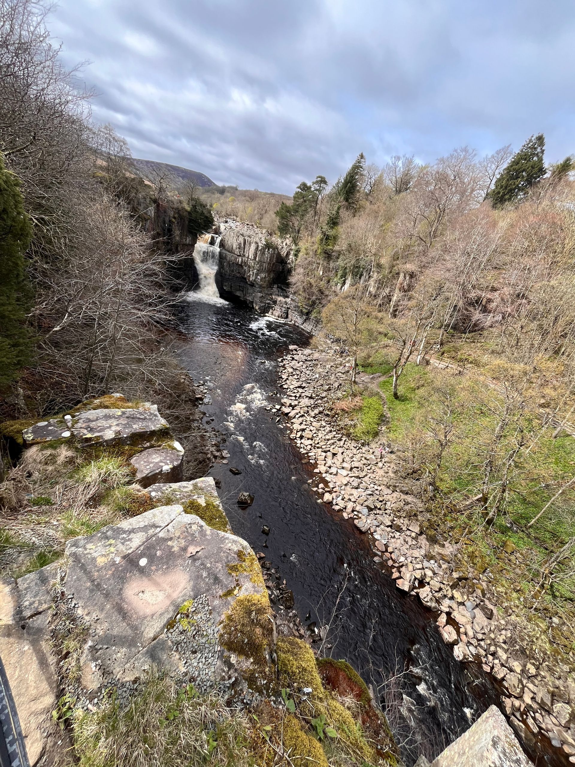 A narrow river flows toward a small waterfall between rocky, brush-covered banks under a cloudy sky.