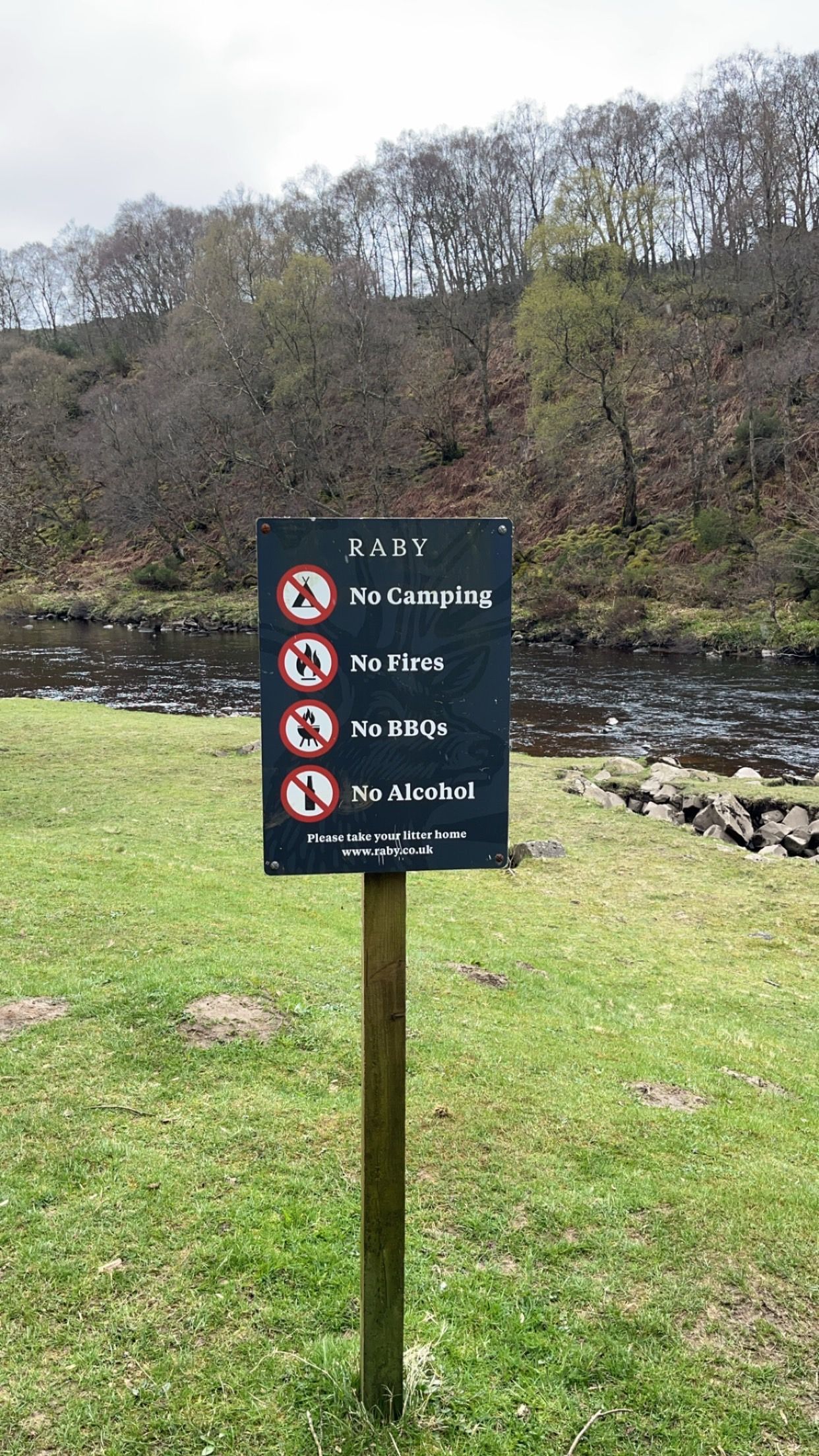 A square sign on a post in a grassy park by a river, listing prohibited activities including camping and fires.