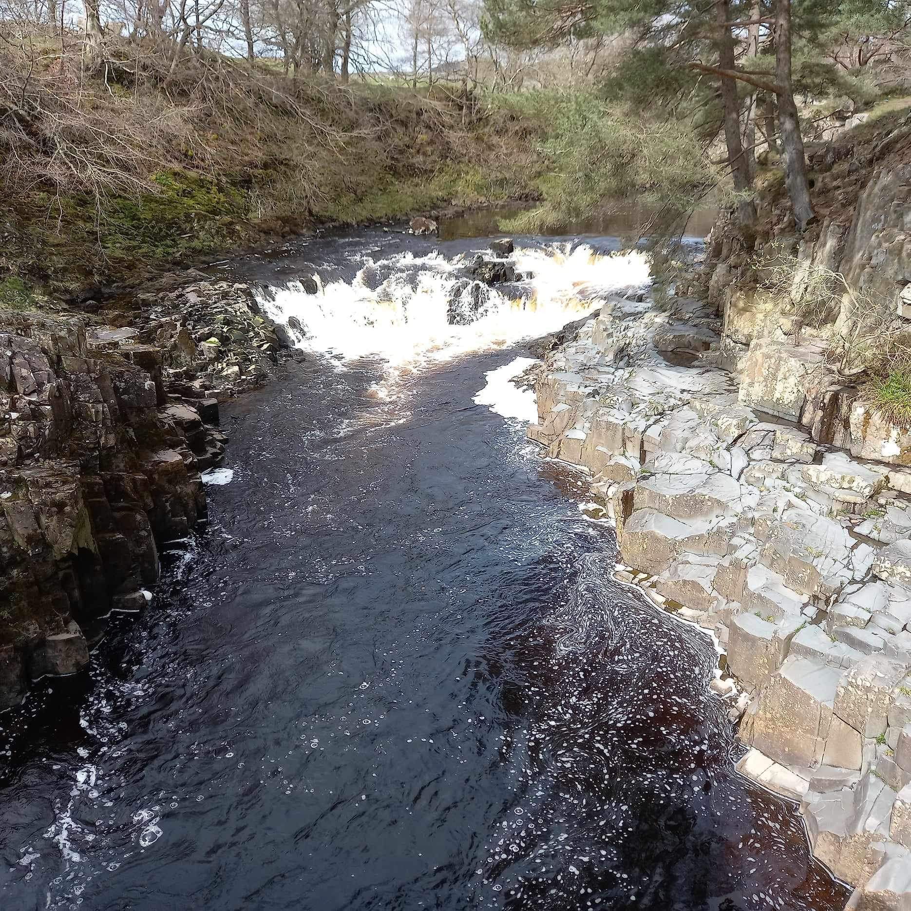 A small waterfall flows into a dark river channel flanked by craggy rock banks and sparse trees.