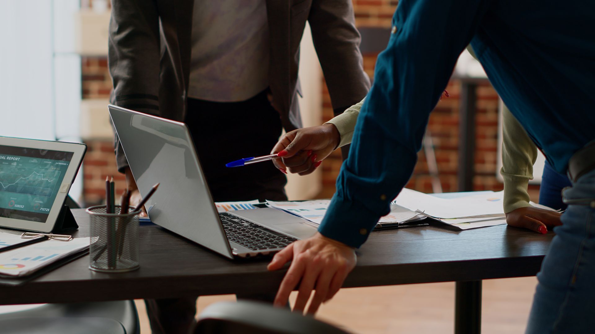 People around a desk looking at a laptop and documents, one pointing with a pen, office setting.
