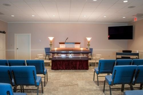 Empty chapel or memorial room with rows of blue chairs facing a lit altar and a wall-mounted screen