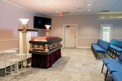 Funeral chapel with an open casket, rows of blue chairs, and a standing lamp in a beige room.
