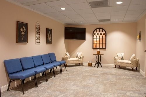 Waiting room with blue chairs, beige sofas, wall art, and a small table beneath an arched window.