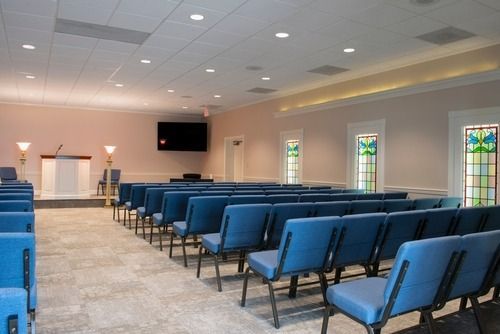 Empty chapel with rows of blue chairs facing a front wall and stained-glass windows.