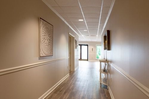 Bright hallway with wood floors, beige walls, framed art, and sunlight at the far end.