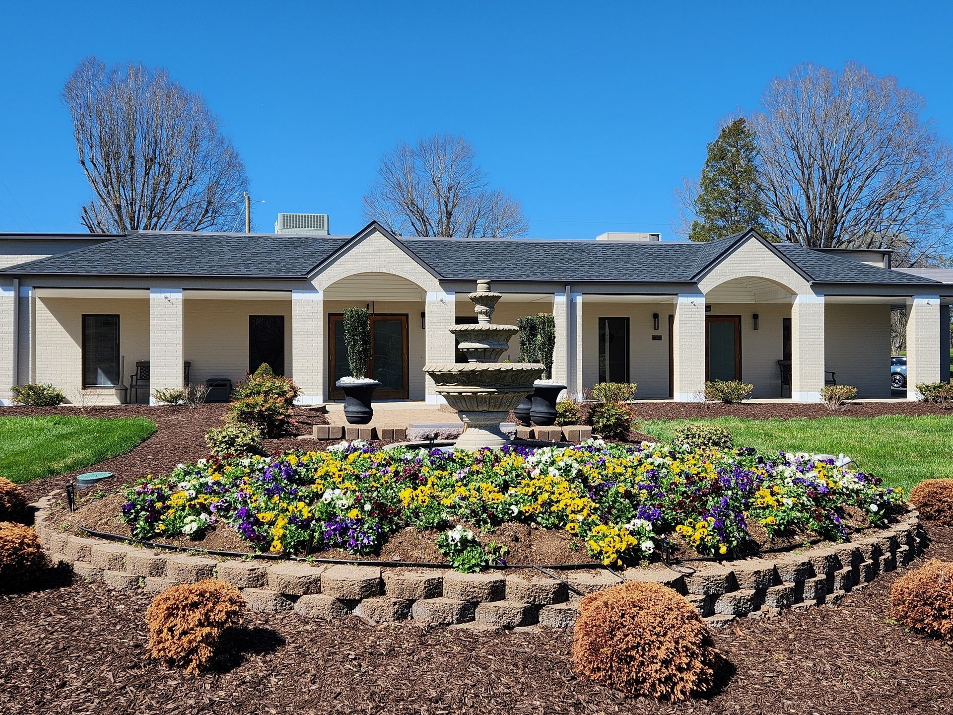 Single-story house with a flower-lined front garden and stone planter under a bright blue sky