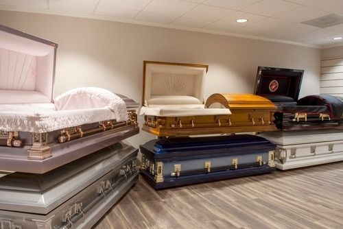 Rows of open caskets in a showroom, including white, gold, blue, and black finishes.