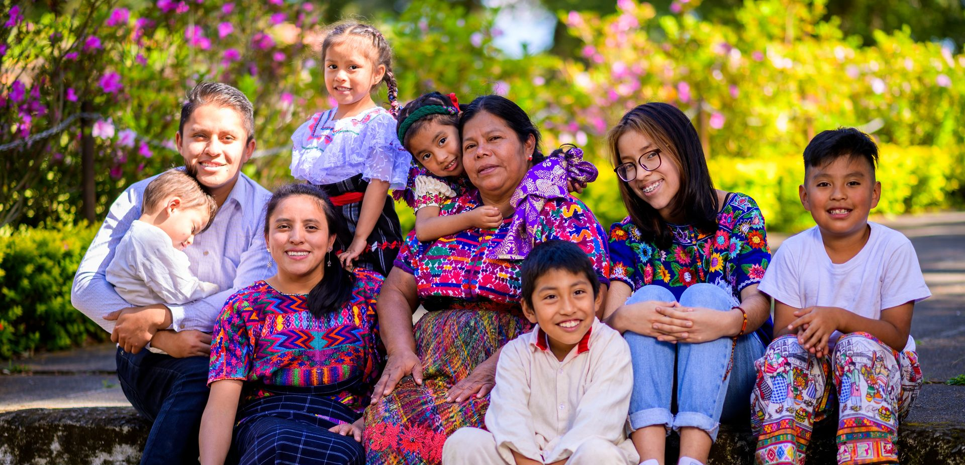 Group posed on steps outdoors, wearing colorful floral outfits, with greenery and flowers behind them