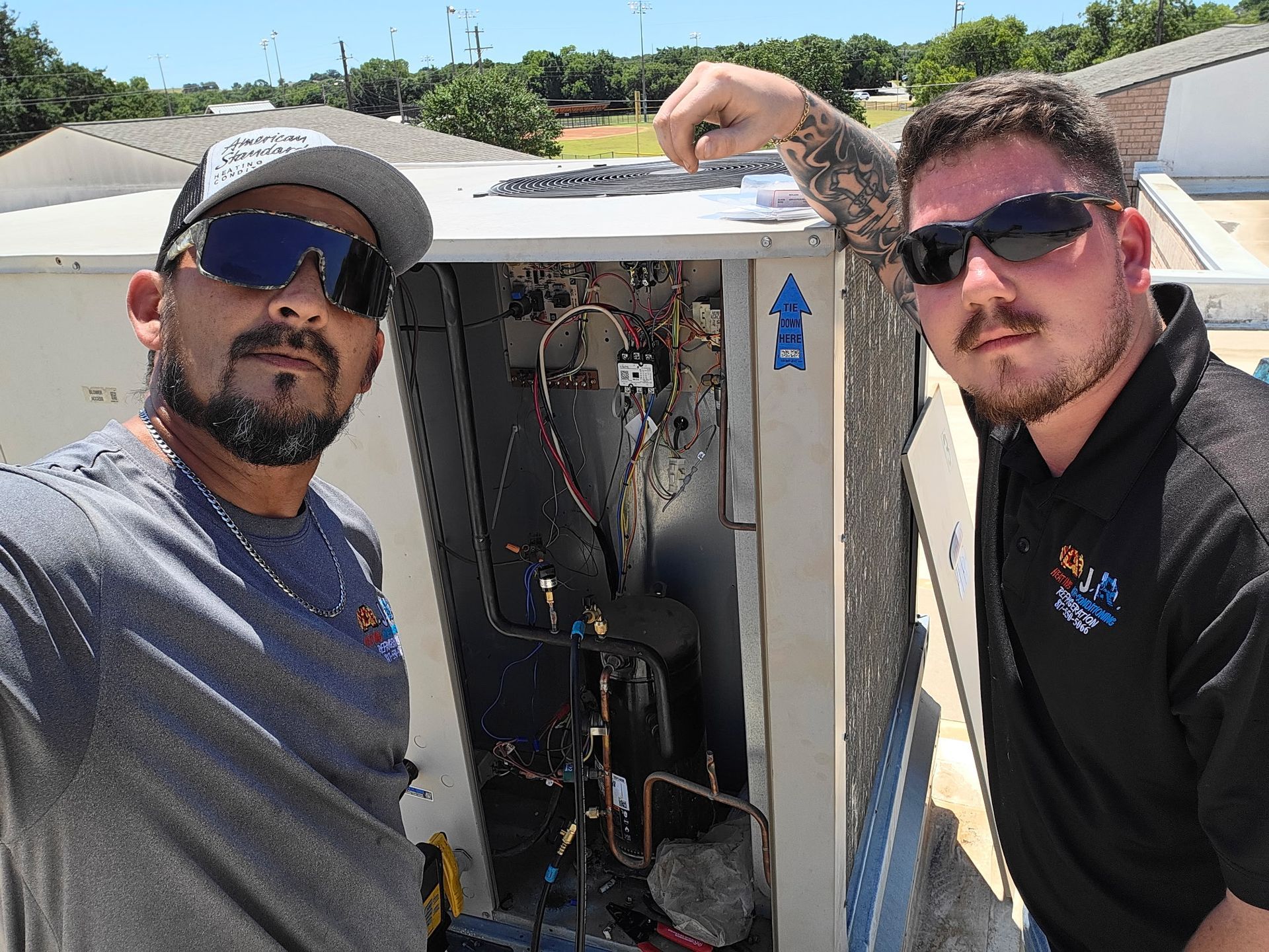 Two men posing beside an open utility cabinet with exposed wiring on a rooftop under bright sunlight