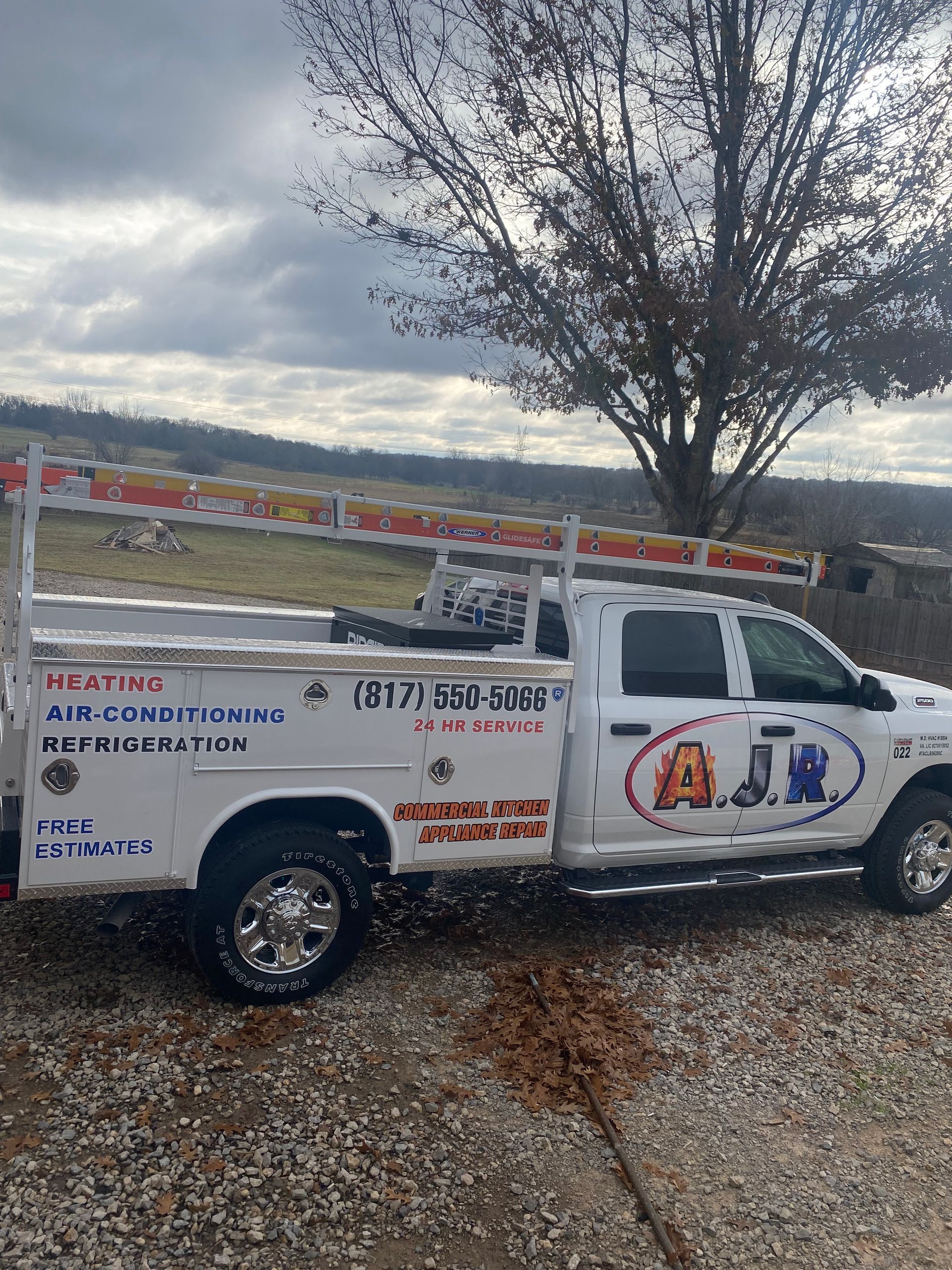 White utility truck with business logos parked on a gravel lot beside a tree under cloudy skies.