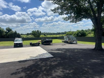 Parked vehicles and trailers on a gravel lot under a tree, with open fields and a cloudy sky beyond.
