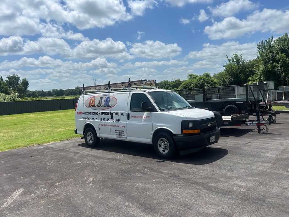 White work van towing a trailer parked on a paved lot under a cloudy blue sky.