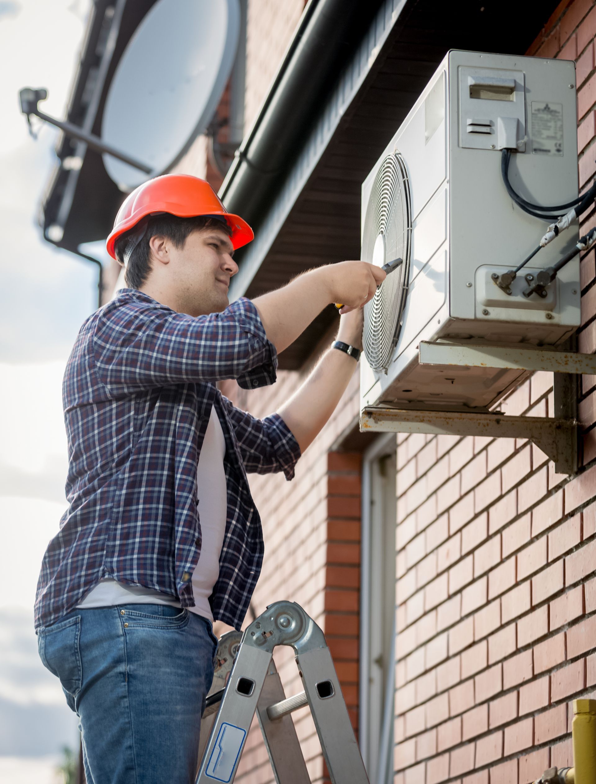 Technician on a ladder repairing an outdoor air conditioner unit beside a brick wall.