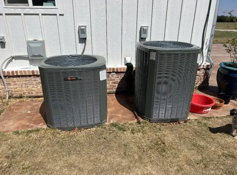 Two outdoor HVAC units beside a white house on a grassy yard.