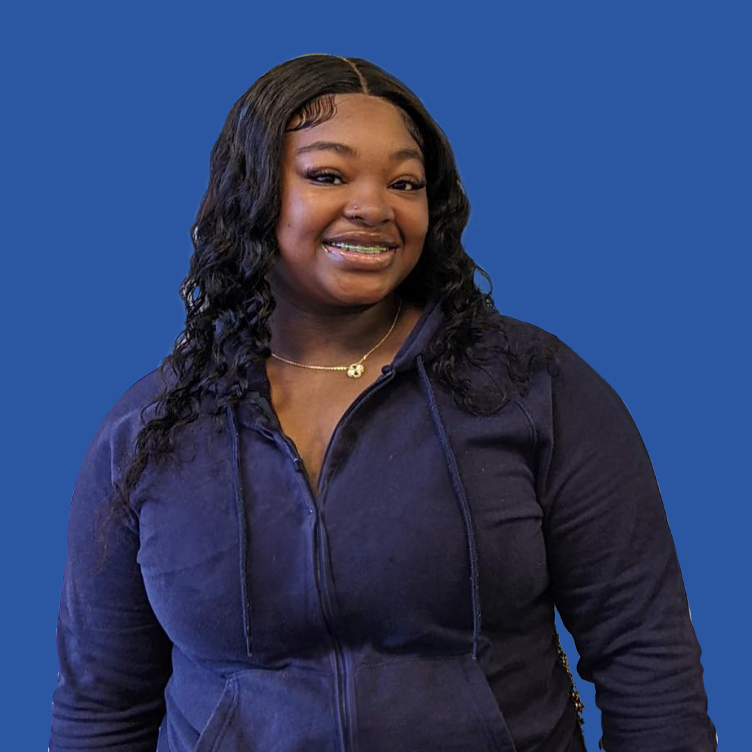 A woman wearing a purple hoodie and a necklace is standing in front of a blue background.