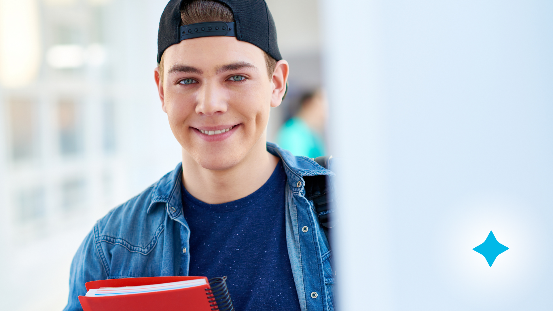 A young man in a baseball cap is holding a notebook and smiling.