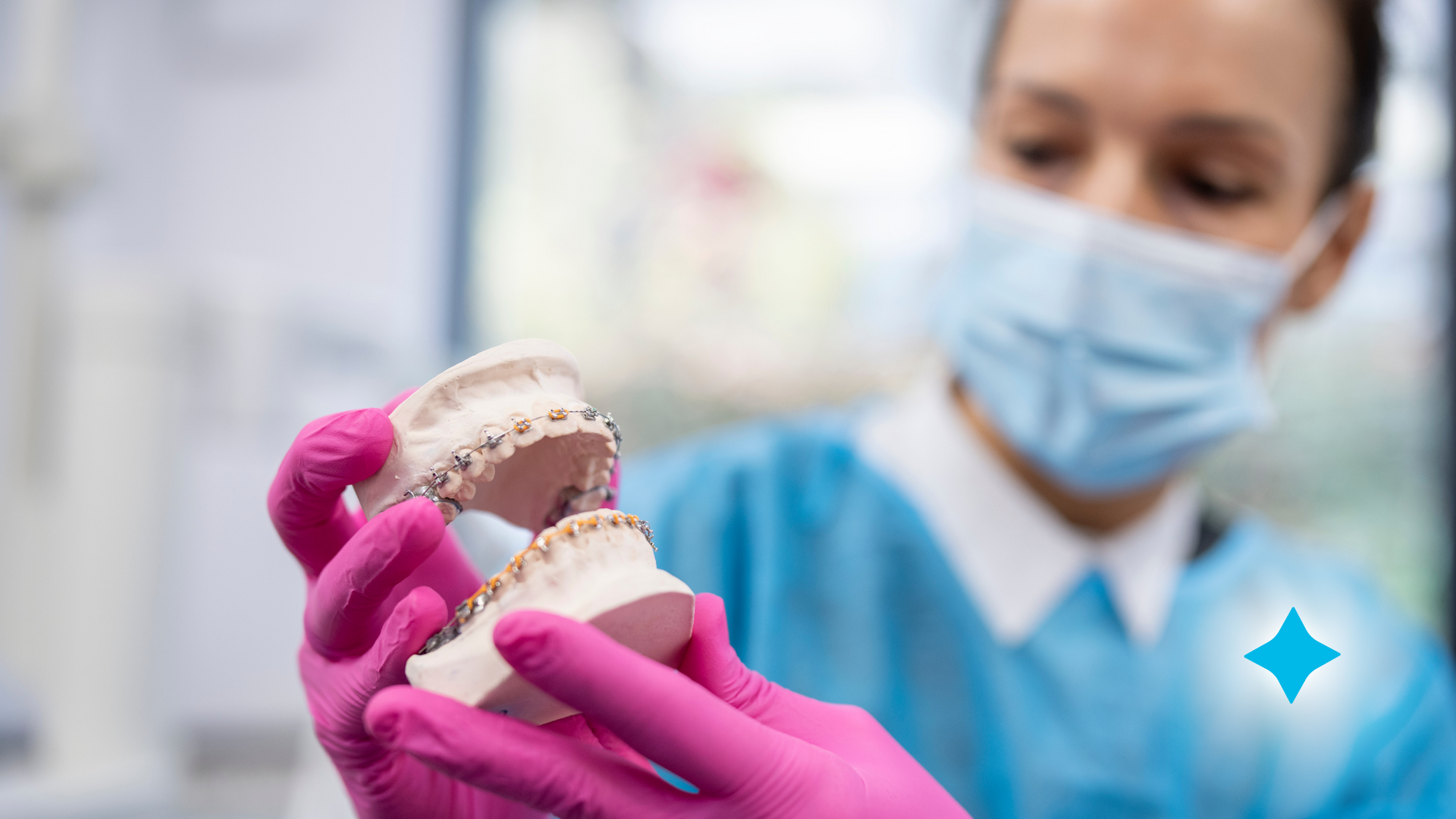 A dentist wearing a mask and pink gloves is holding a model of teeth with braces.