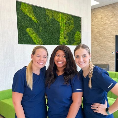 Three women in scrubs are posing for a picture in front of a green couch.