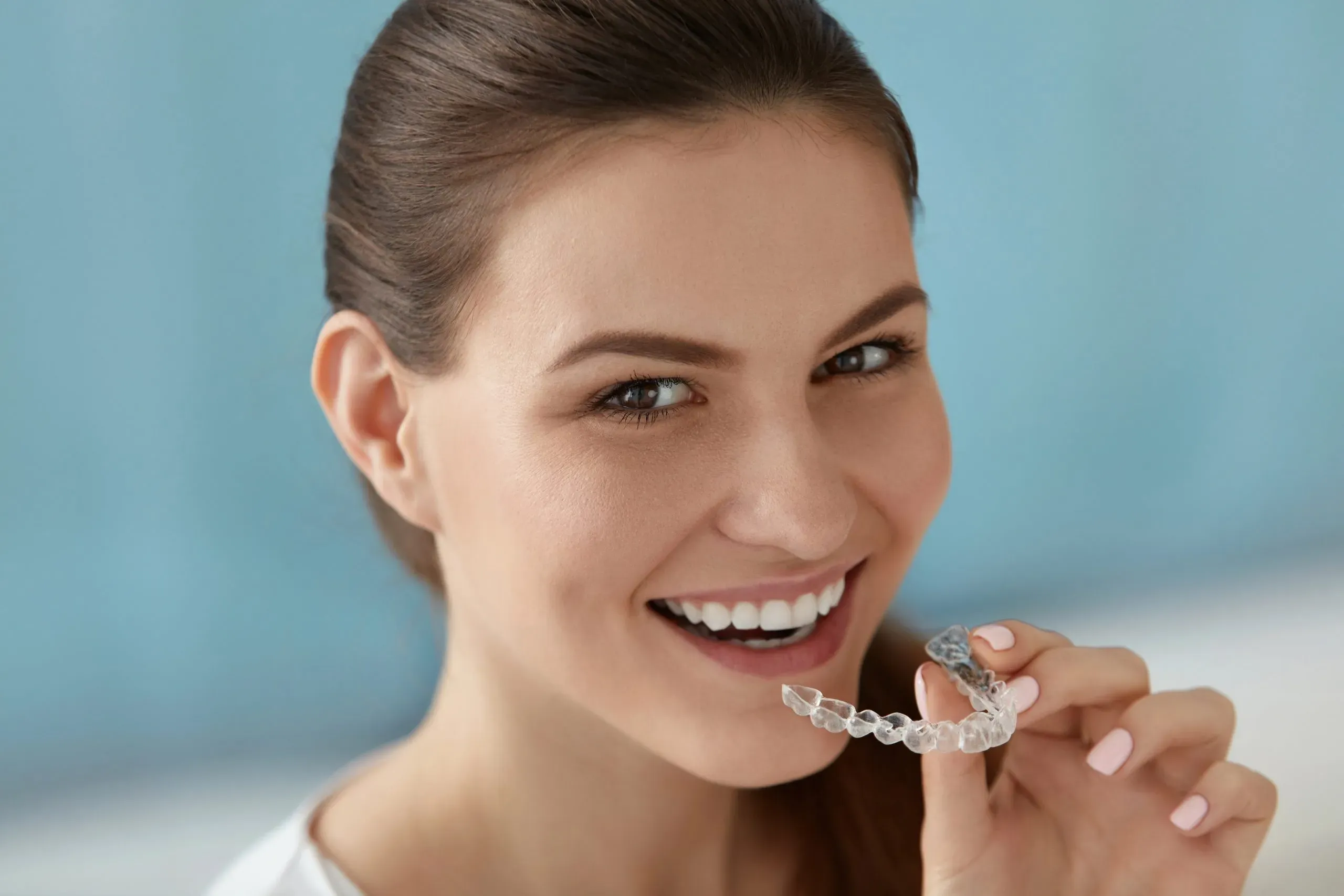 A woman is smiling while holding a clear retainer in her mouth.