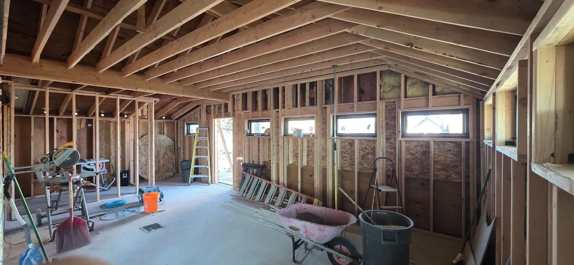 Unfinished attic with exposed wooden framing, insulation, windows, and construction tools scattered on the floor.