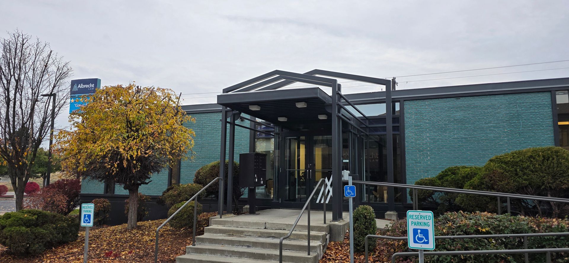 Building entrance with steps, glass doors, covered awning, and accessible parking sign on a rainy day