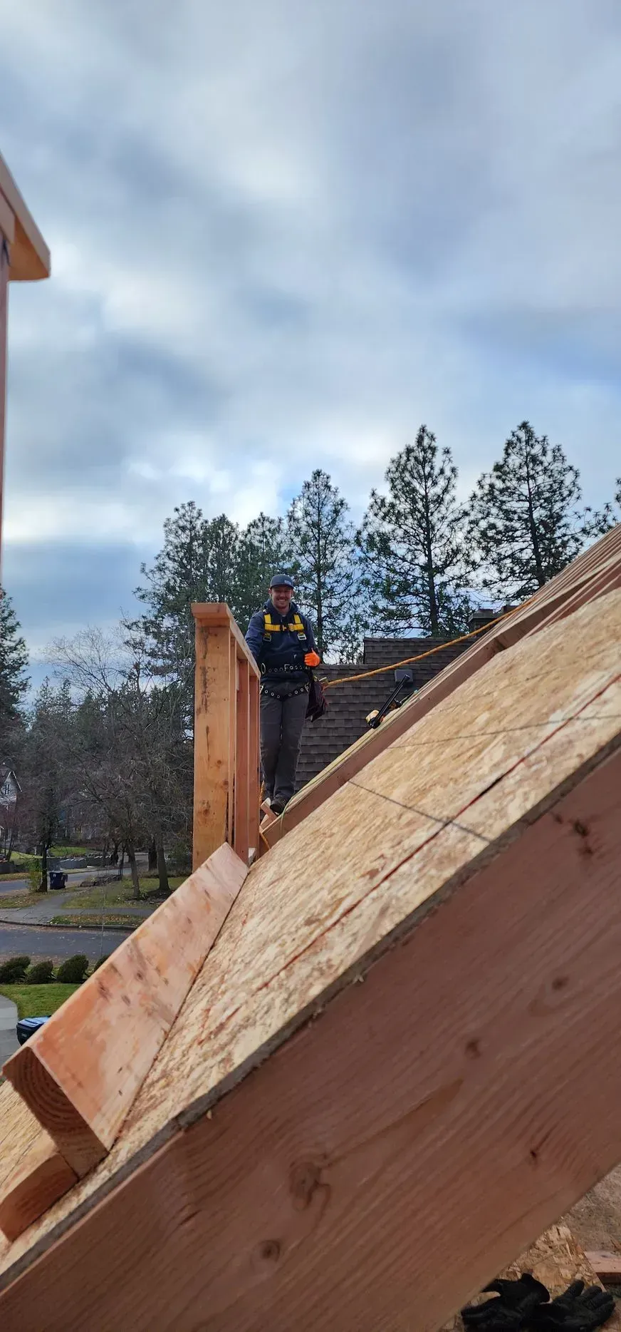 Person in a blue jacket stands on a wooden roof frame outdoors under a cloudy sky.