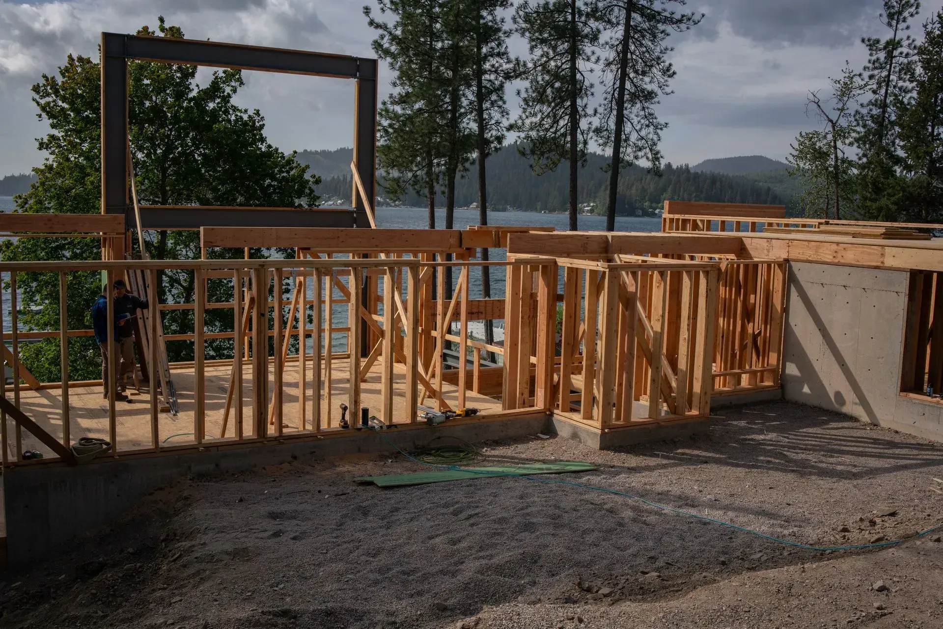 Wood-framed building under construction on a gravel lot with a lake and trees in the background