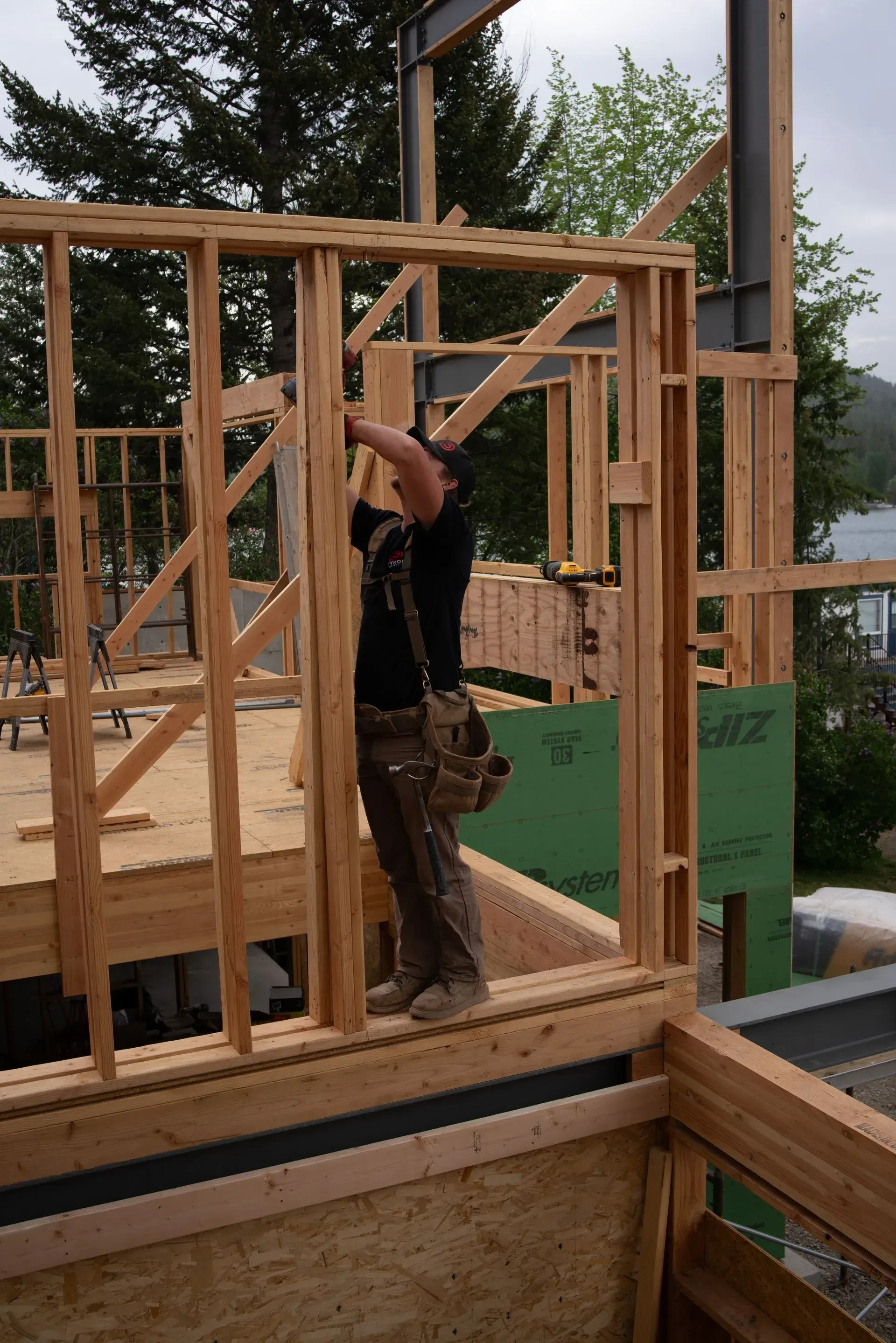 Person framing a wooden house wall at a construction site, with exposed studs and plywood sheathing.
