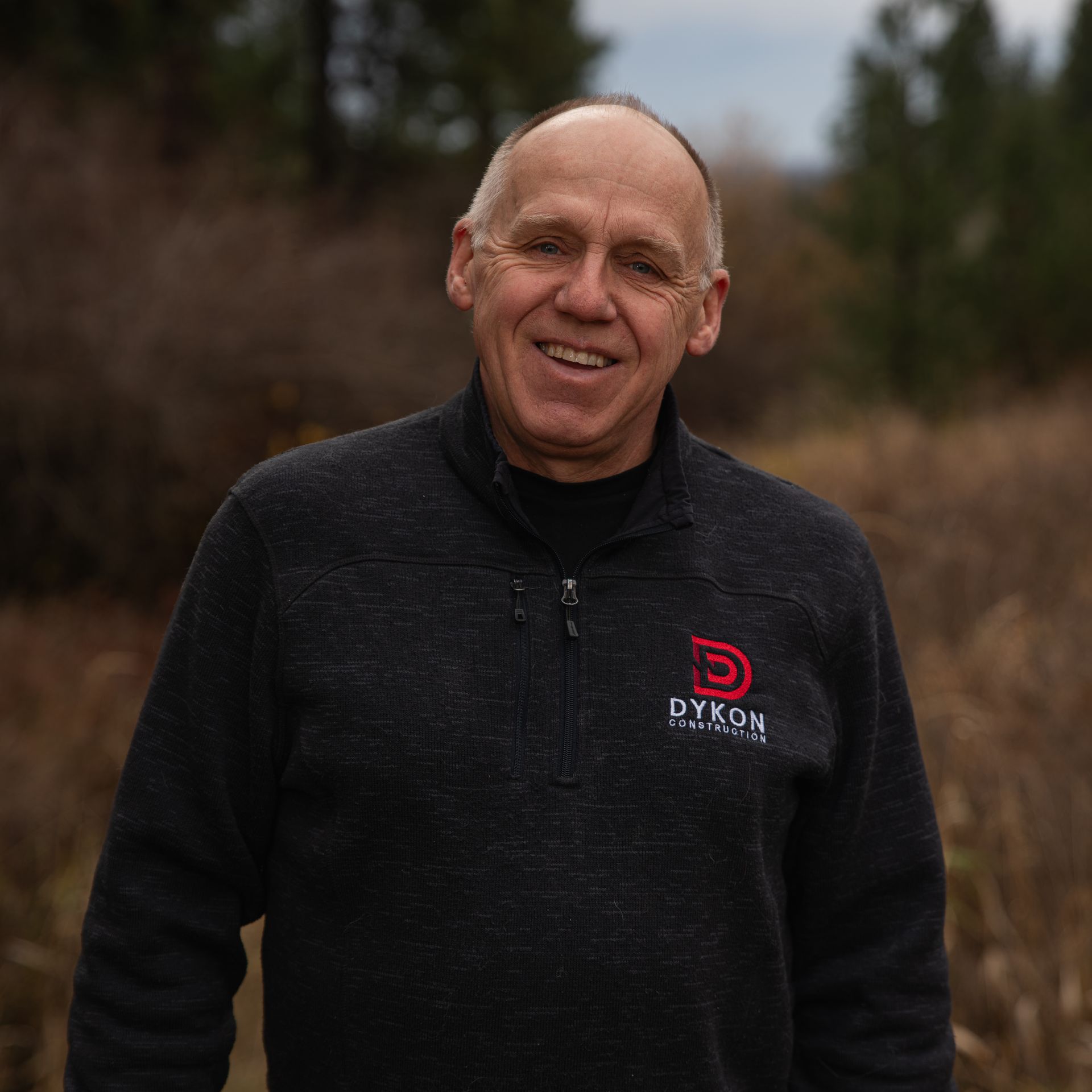 Smiling man in a black fleece jacket with a red logo, standing outdoors in a blurred wooded area.