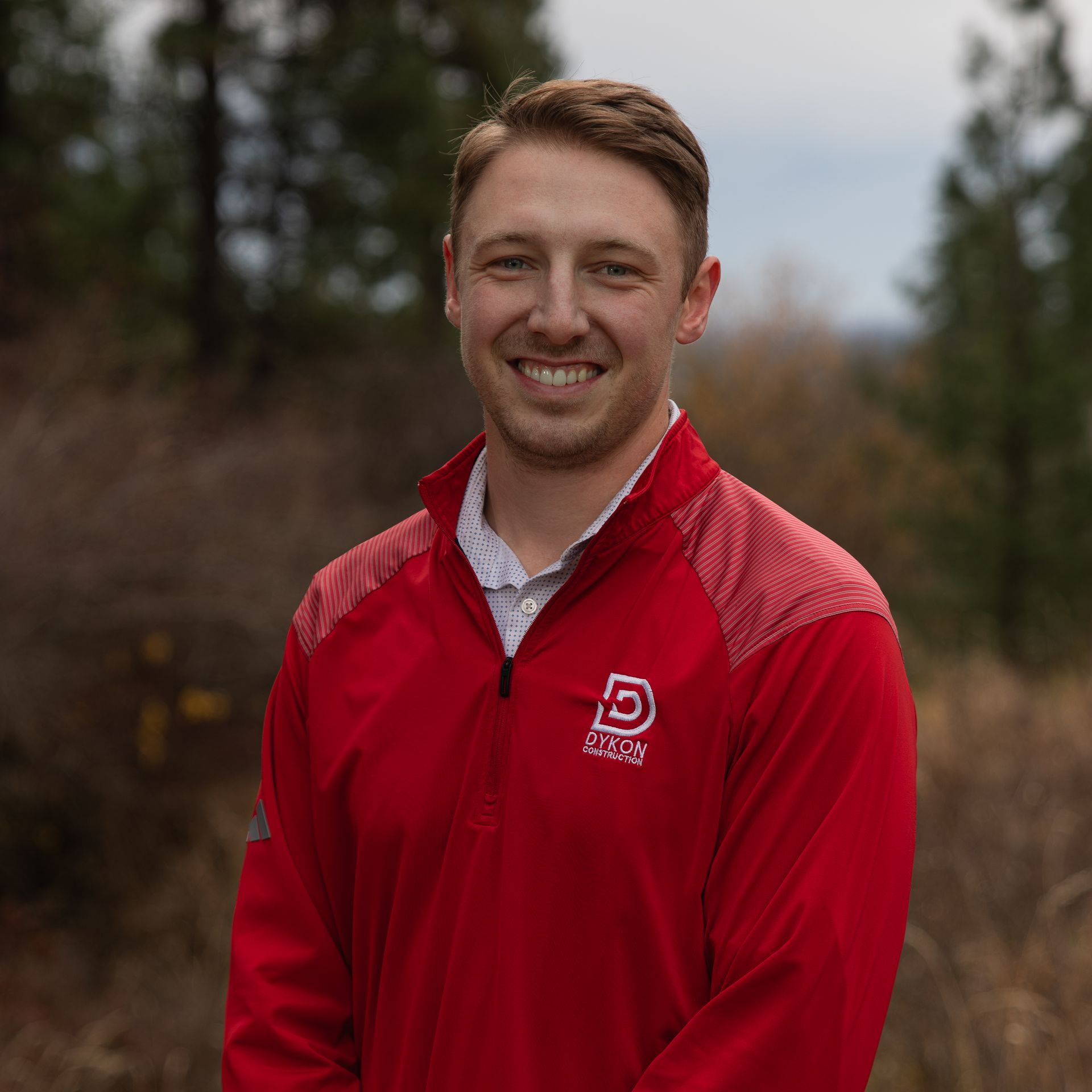 Smiling man in a red jacket standing outdoors on a blurred wooded path