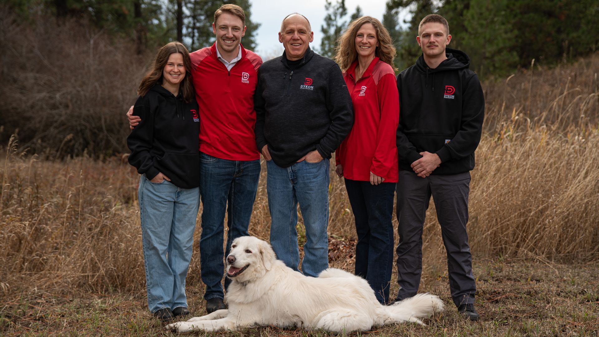 Five people in red and black jackets pose outdoors with a white dog lying in front.