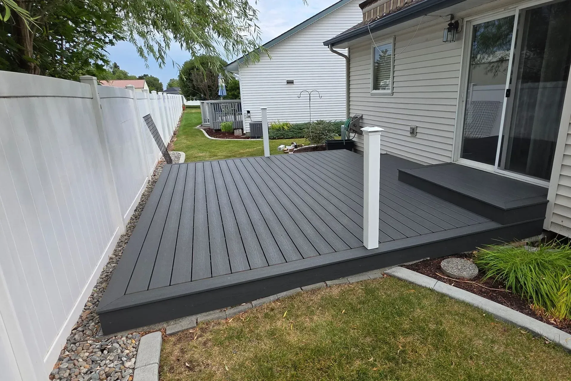 Small gray composite deck beside a house, bordered by a white fence and grass yard.