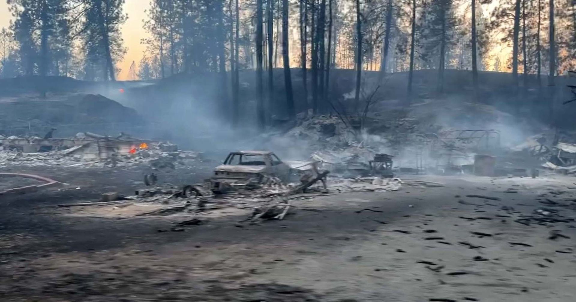Charred debris and a burned-out car amid smoke in a scorched forest clearing